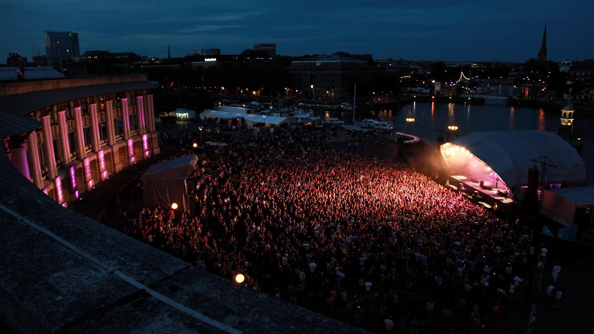 A night time shot of Lloyds Amphitheatre in Bristol. The main stage is covered with a canopy. Thousands of fans are standing in front of it watching a gig. The silhouette of the Bristol skyline can be seen in the distance 