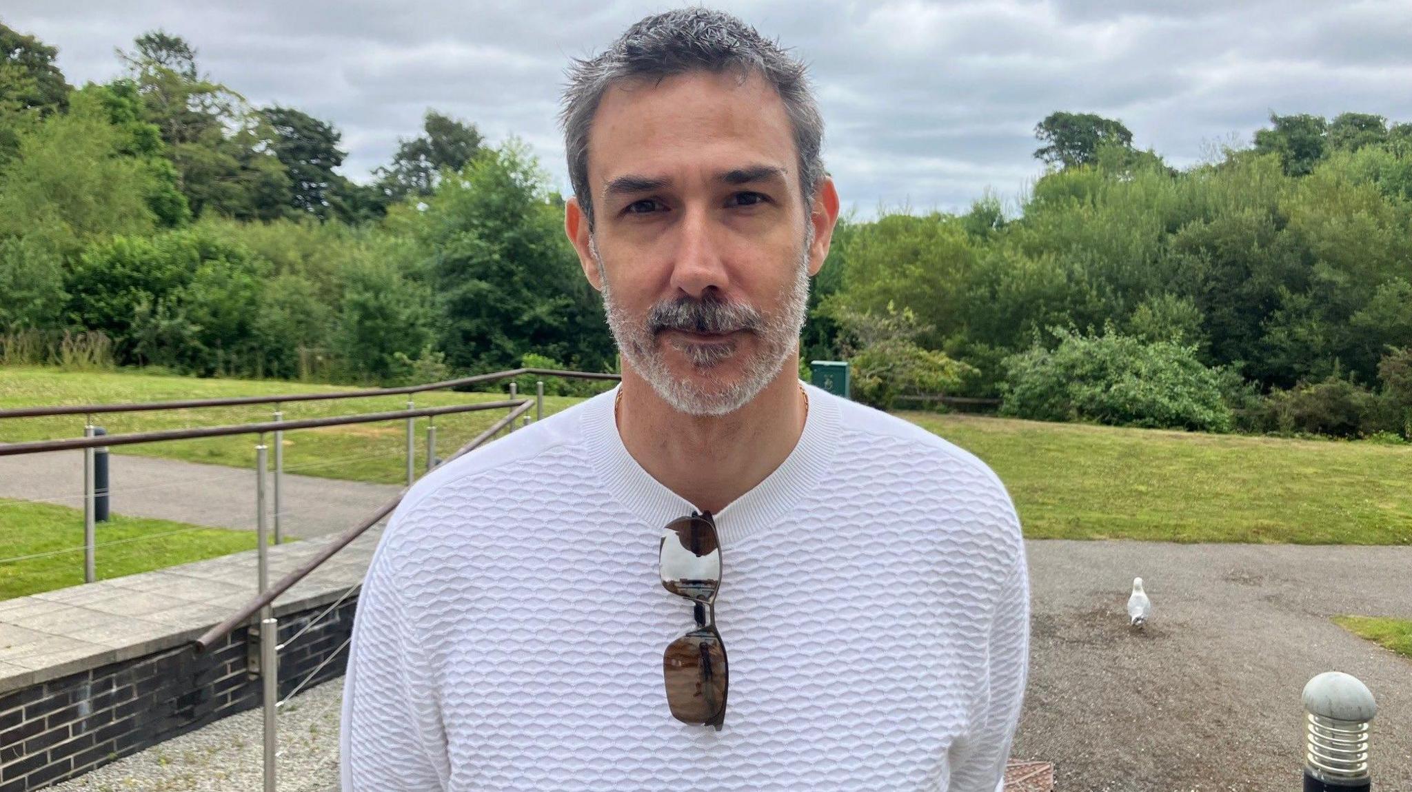 A man with a grey beard smiles, He's standing in front of woodland at Falmouth university's Penryn campus 