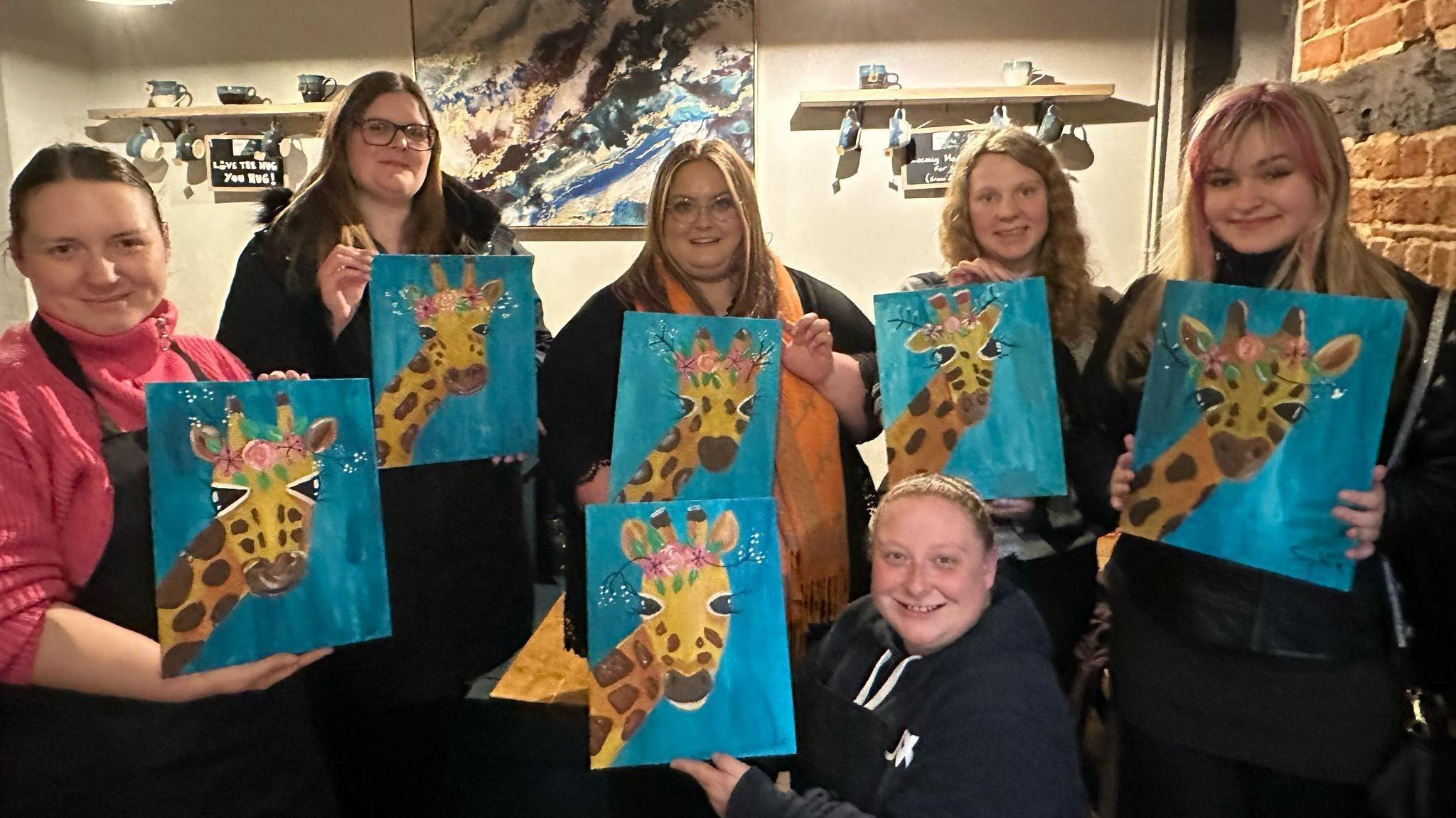 Women smile at the camera while holding paintings they had all done of a giraffe on a blue background.