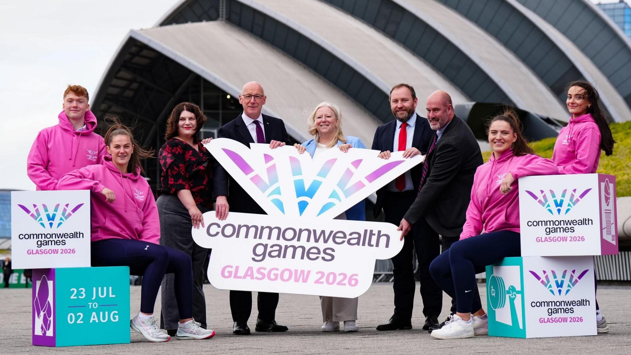 Athletes in pink hoodies flank official figures who are standing outside the SEC Armadillo in Glasgow holding Commonwealth Games branding. They are, from left, Glasgow City Council leader Susan Aitken; First Minister John Swinney; Commonwealth Games Federation chief executive Katie Sadleir; Scottish Secretary Ian Murray and the chief executive of Commonwealth Games Scotland Jon Doig.