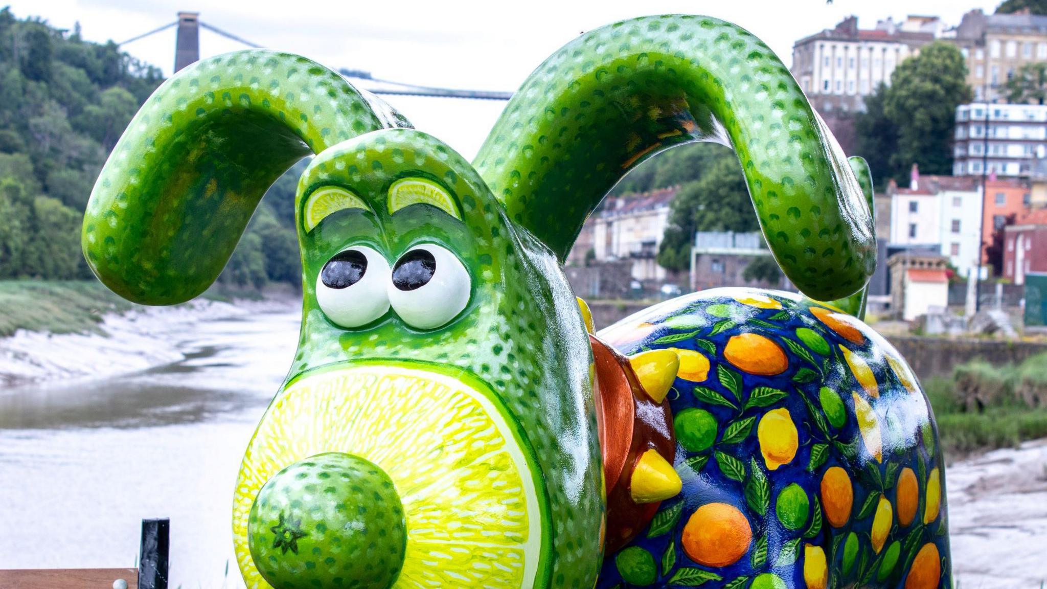 A close-in view of a Gromit structure that has been painted in bright colours and has a green and yellow nose. In the background the Clifton Suspension Bridge is visible