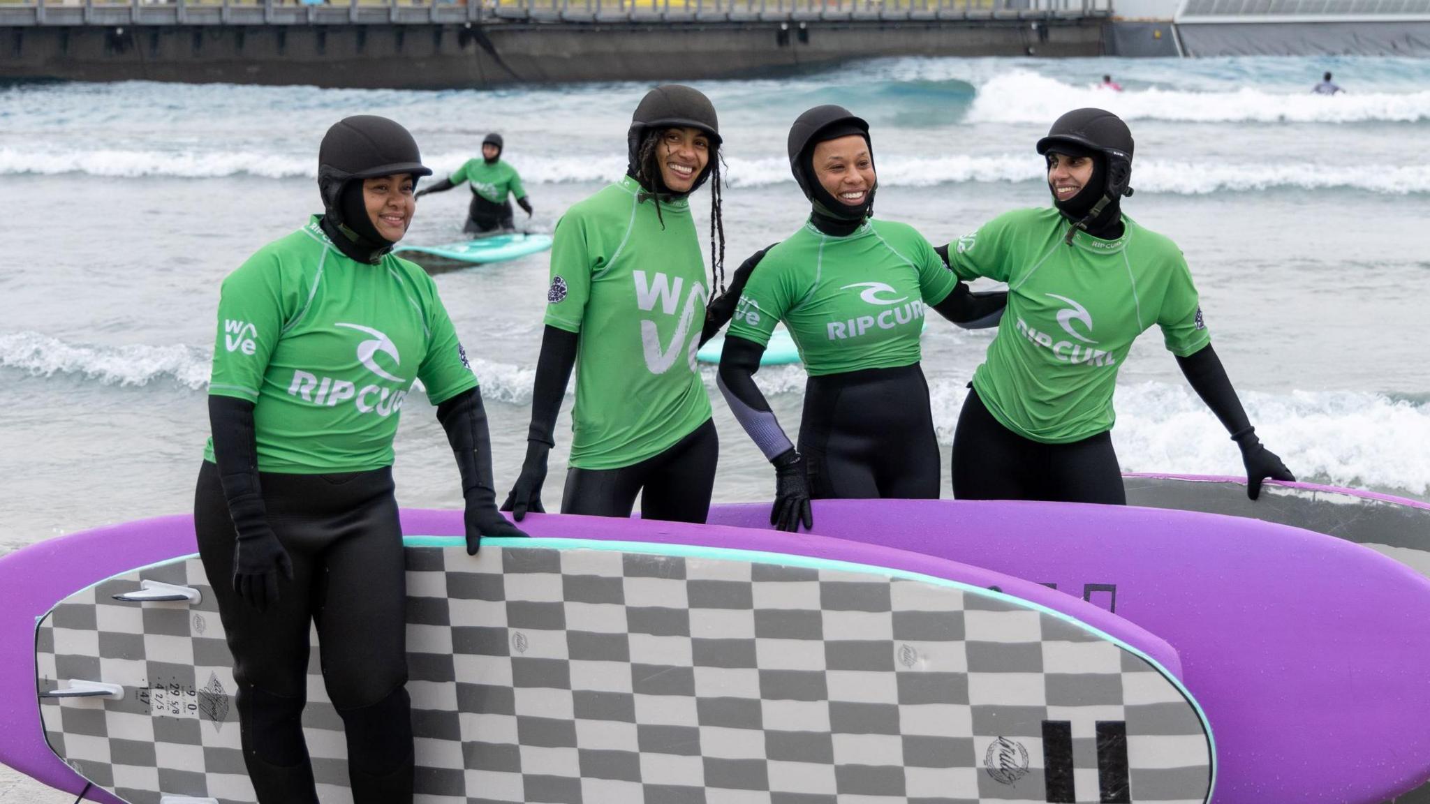 Four young women in wetsuits and protective hats stand smiling at the camera with their surfboards at The Wave in Bristol. Other surfers can be seen in the water in the background