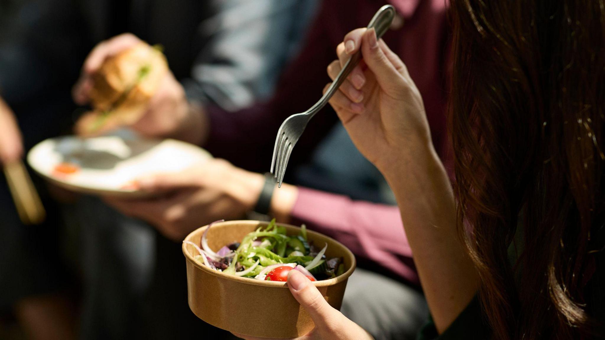 A woman's hands holding a fork and a cardboard takeaway pot with a salad in it. Her long dark hair can be seen on the right hand side of the frame, and a man eating a sandwich can be seen in soft focus behind her.