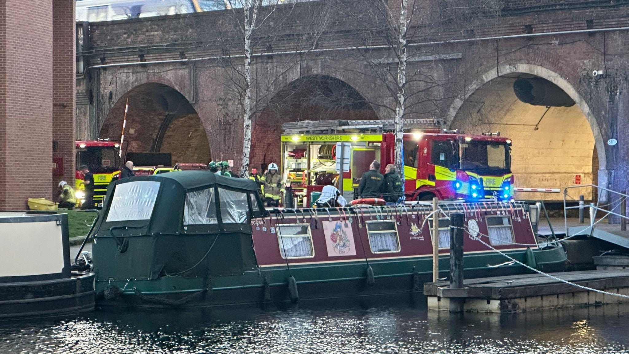 A green and burgundy coloured barge boat docked on the water. In the background there is a fire engine with its lights illuminated. It is parked in front of a bridge with three arches in it. 