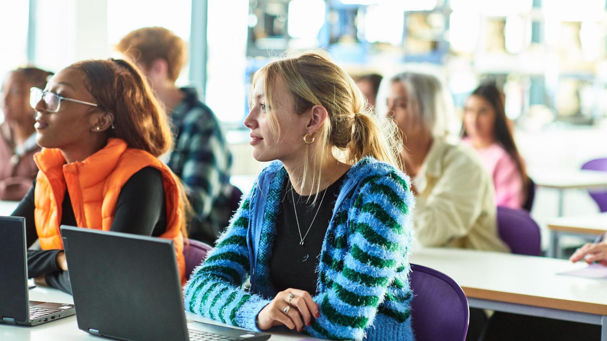 Female caucasian student with blonde hair sitting in class in front of an open laptop at sixth form college listening and paying attention.