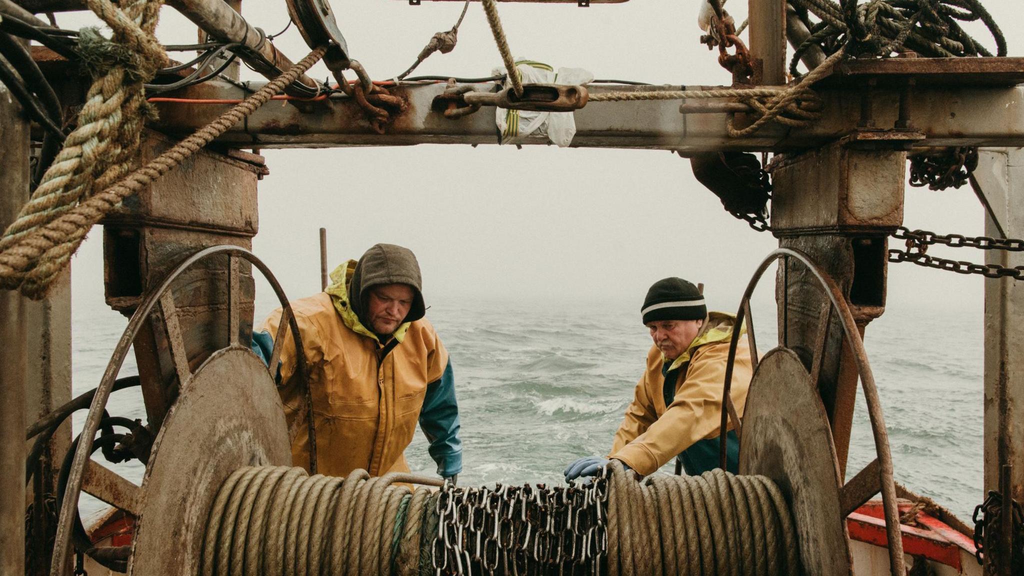 A coloured image of a father and son on the back of a fishing boat. They are both wearing yellow waterproofs and protecting their heads from the rain. Behind the men, mist can be seen on the top sea.  The men are looking down as the pull in heavy chains. 