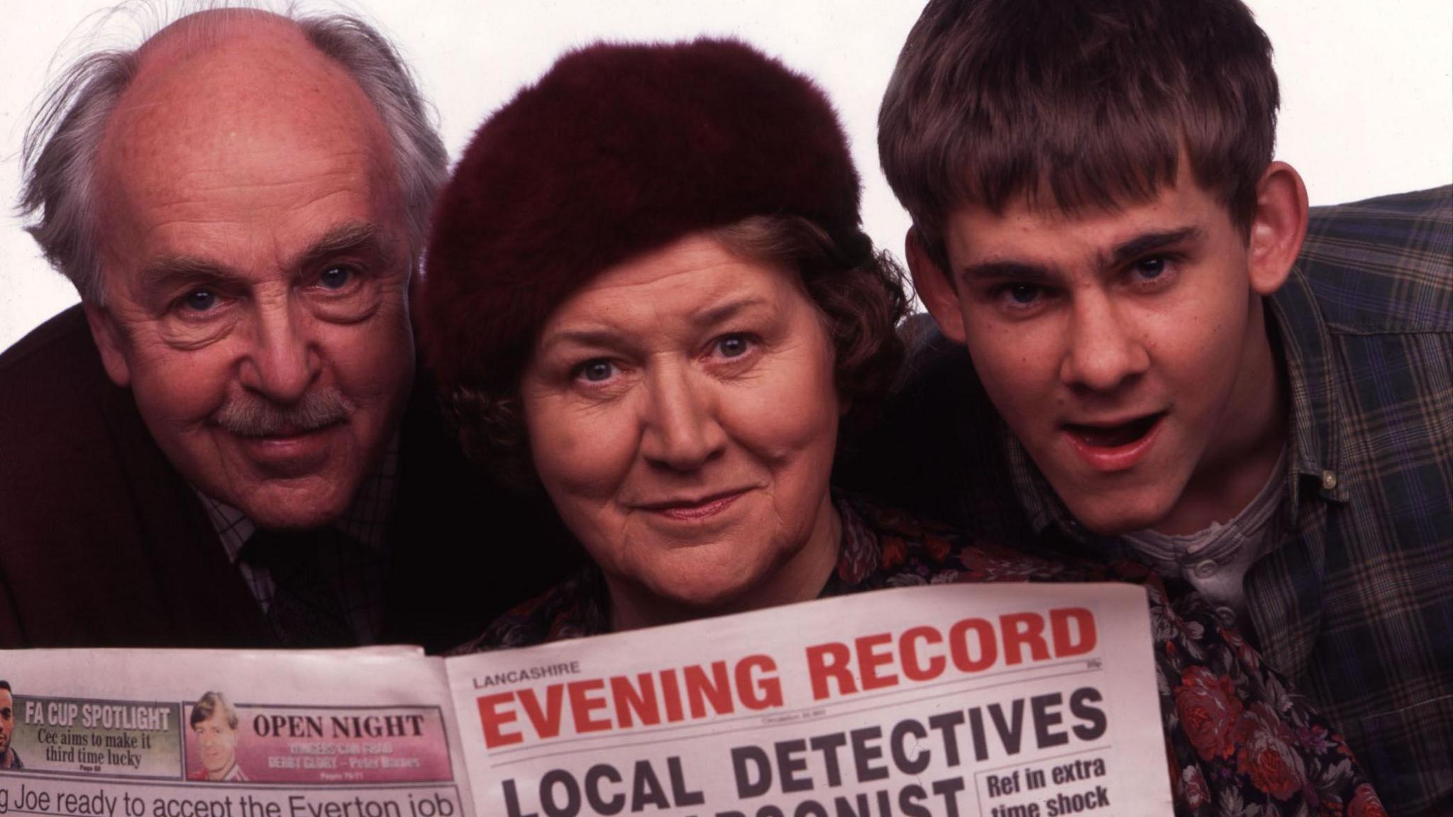 Routledge, Derek Benfield and Dominic Monaghan looking over the top of a Lancashire Evening Record newspaper with a partially-visible headline about "local detectives".
