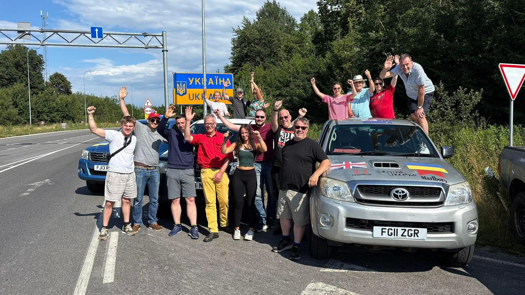 A group of about 20 people standing on the side of a road cheering, with a sign for Ukraine behind them. Alastair Caldwell is pictured with a group of three other people stood at the back of the Toyota Hilux pick-up.