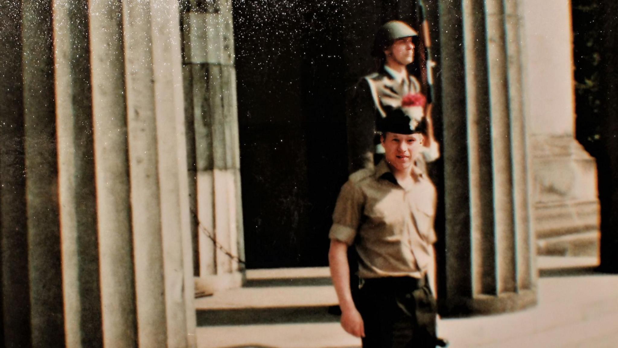 An archive picture of a soldier posing at the camera. He is a beige military uniform shirt with a beret on his head. He is stood in front of a building fronted by large pillars. In the background you can see a guardsman holding a rifle looking away from the camera.