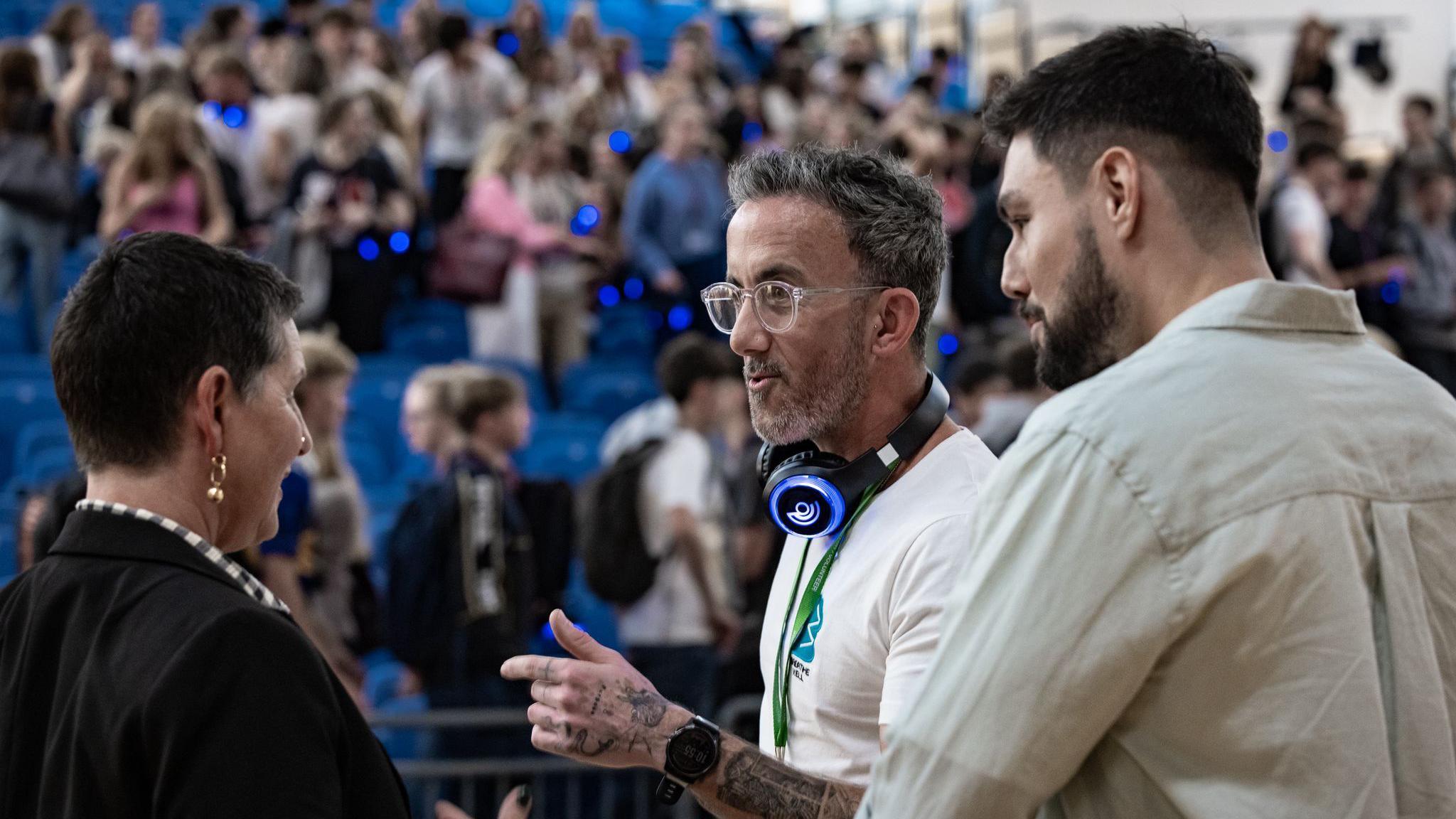 Mick Phipps is standing in front of an audience in what appears to be a school hall. He has headphones around his neck and he is speaking to a dark-haired woman. A dark-haired man is standing next to him.