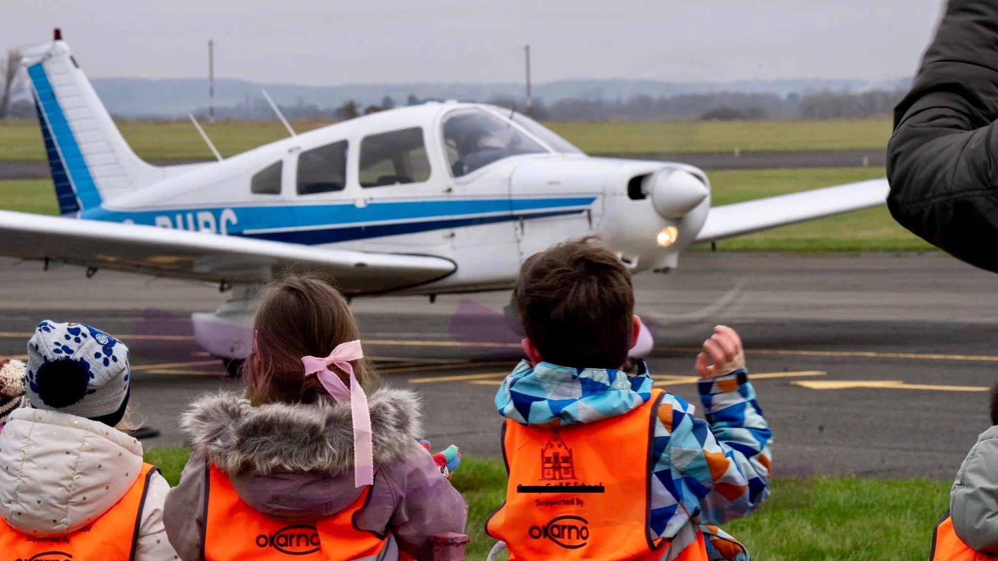 Children in high-vis vests watching a light aeroplane take off