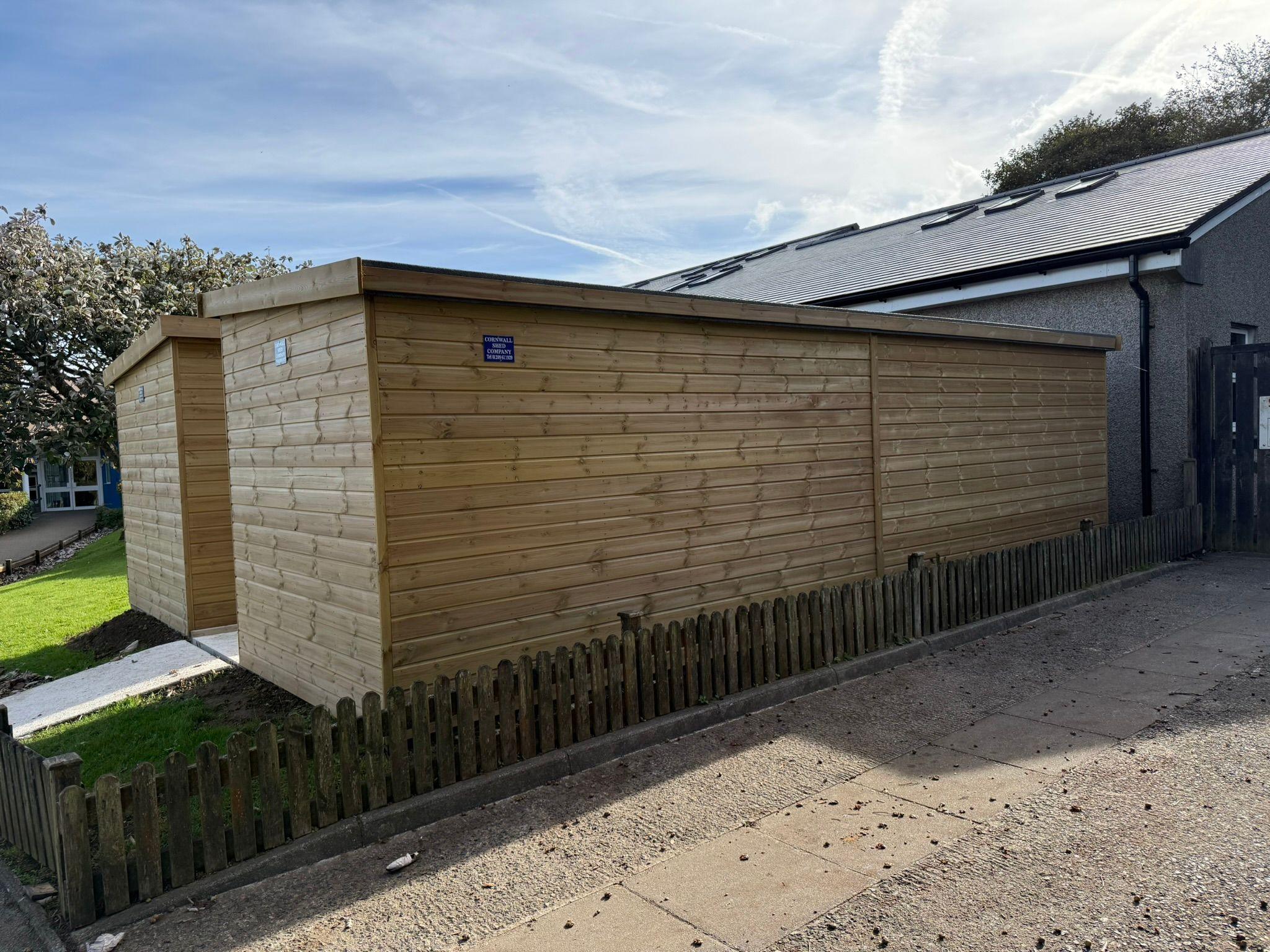 A large wooden shed is installed behind a picket fence on some grass, with a tree behind it. There is a path leading to the shed, and a paved area in the foreground.