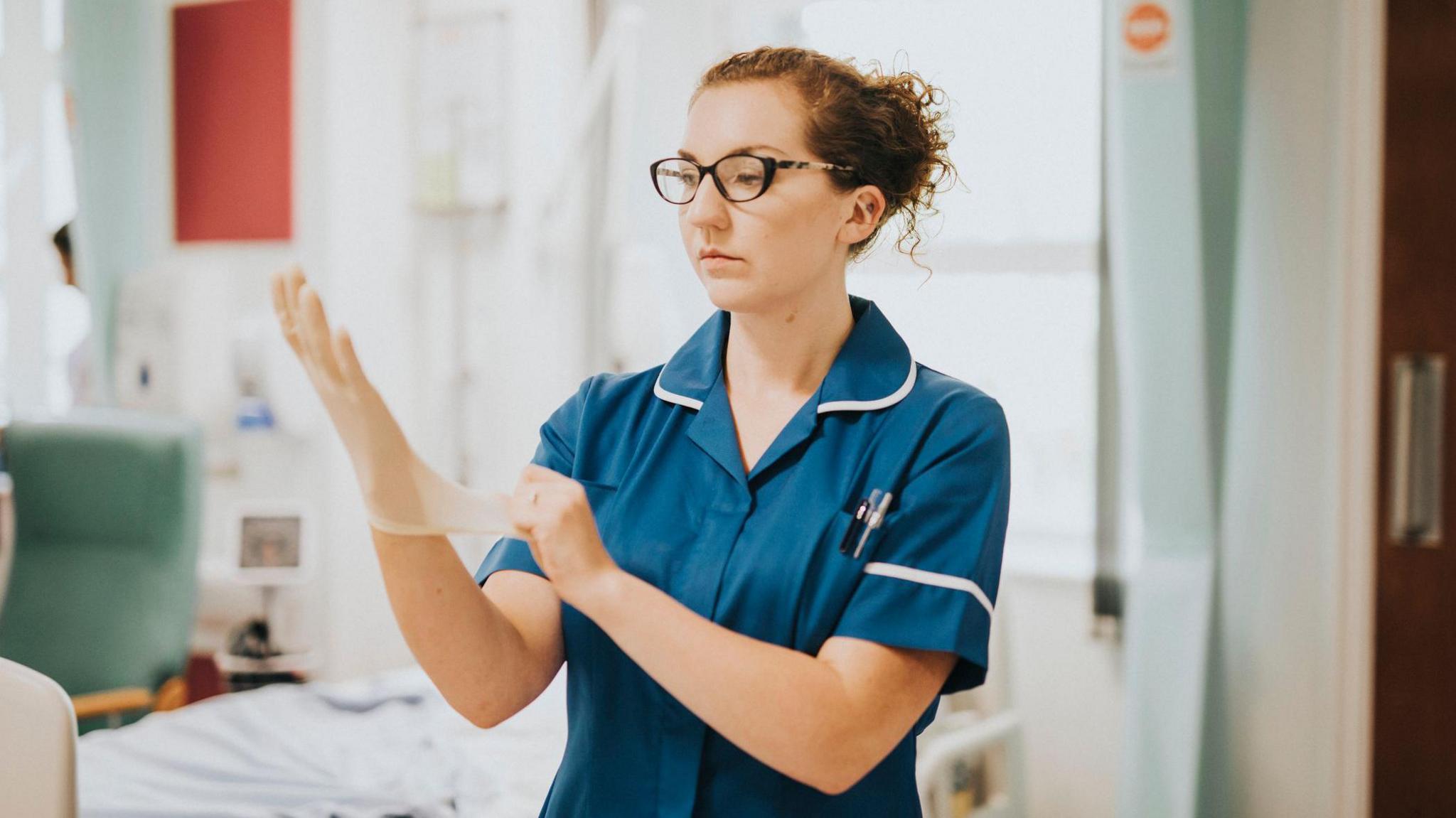 Nurse wearing turquoise uniform applies plastic gloves in a hospital word