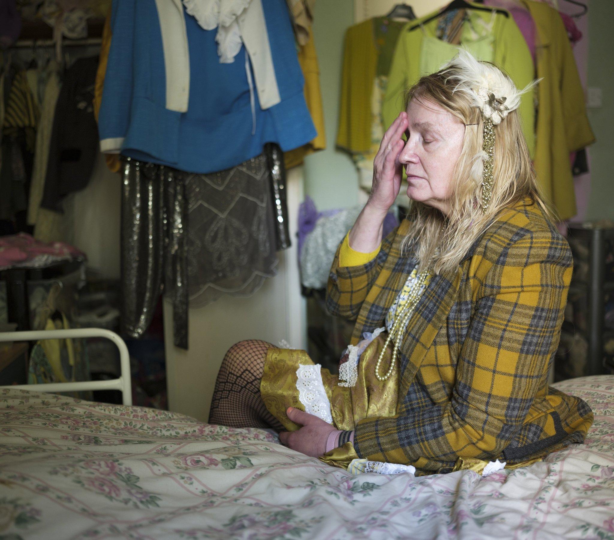 Liz holding her head sitting on her bed with dresses hanging on the walls around her