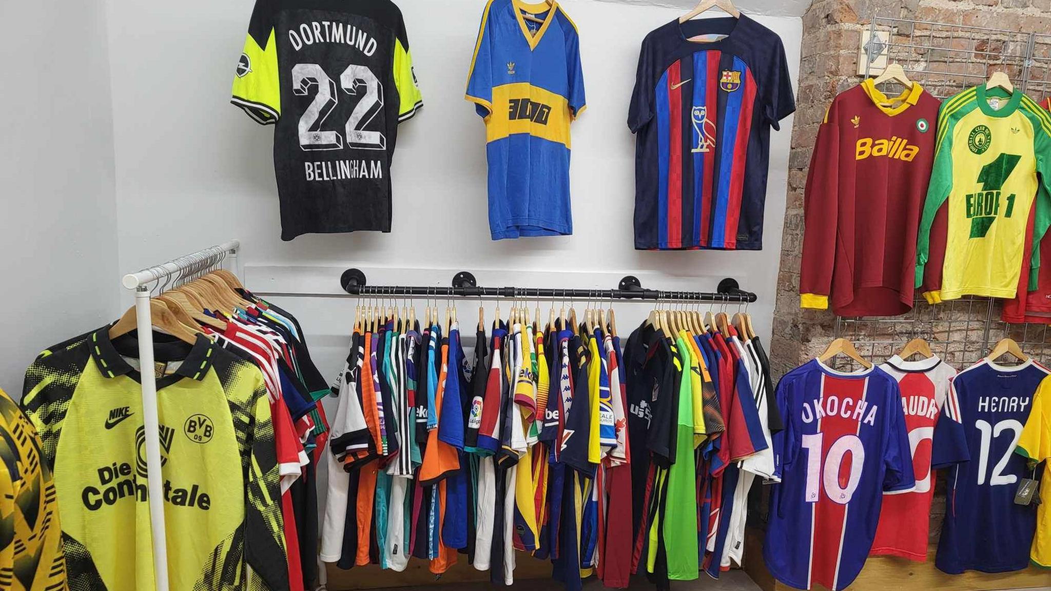 Two rails, on the left and centre, hold dozens of bright retro football shirts. There are three shirts hanging on the centre wall, one reads "Dortmund. Bellingham". To the right, there is a metal grate attached to a brick wall with more shirts hanging up.