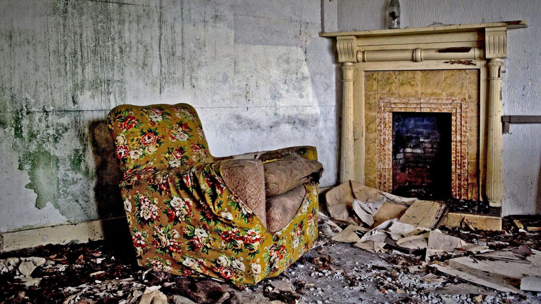 An armchair, its floral patterned cover torn, sits in a derelict room of an abandoned house. There is mould on the wall behind it and a fireplace on adjoining wall. Broken plaster covers the floor.