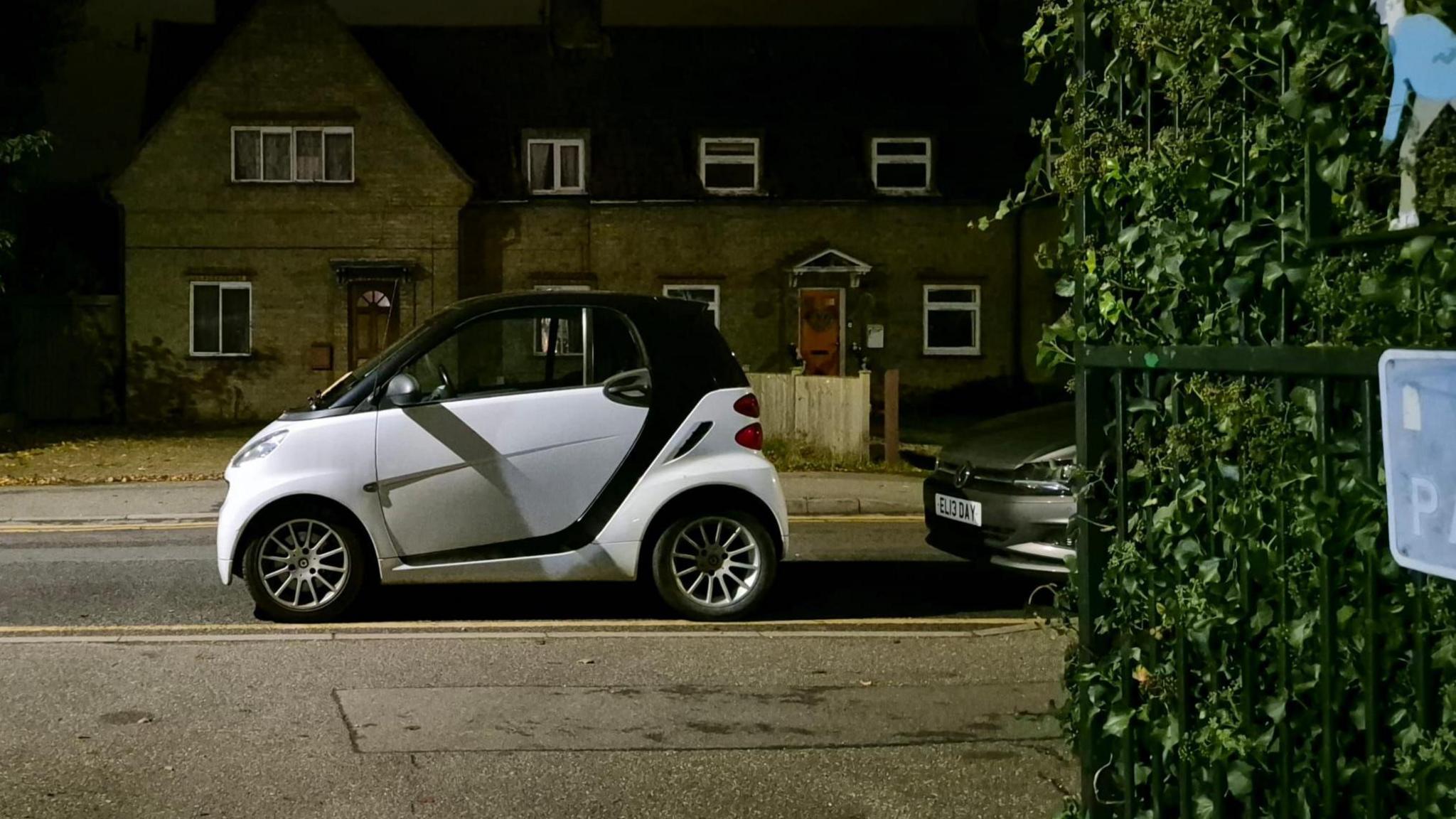 A black and white Smart car is parked across a dropped kerb outside a bowls club. There is another car behind it and houses in the background