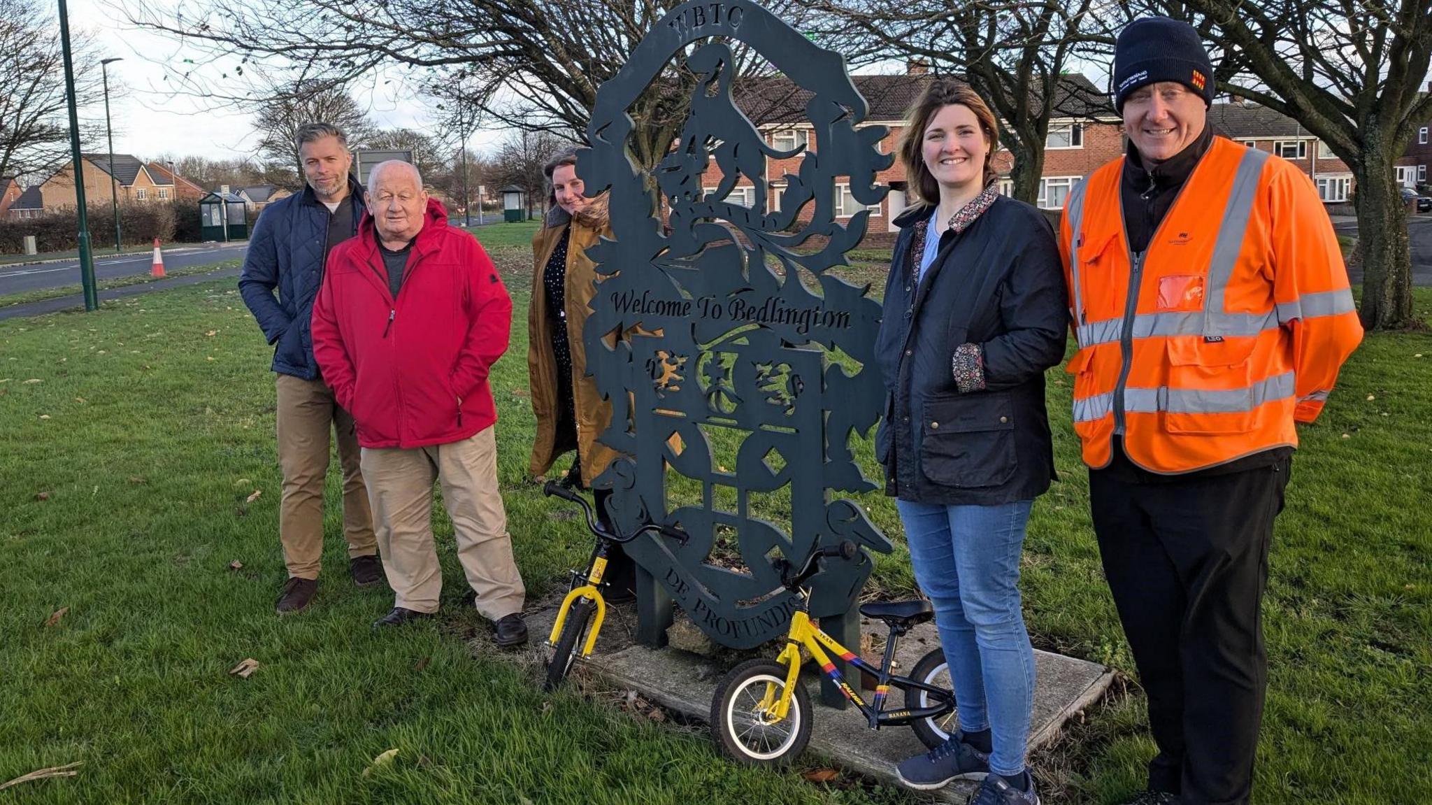 Five people standing around a sign saying Welcome to Bedlington. There are two very small bikes at the base of the sign.