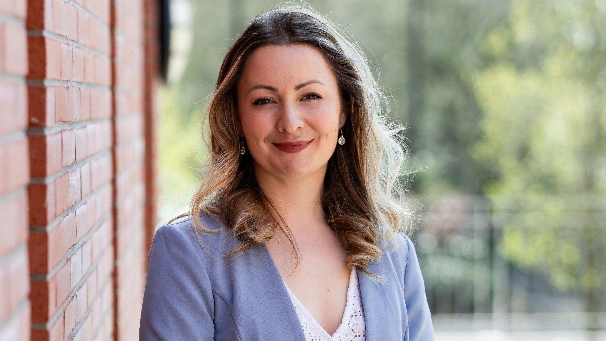 A close-up image of Dr Kathy Ruddy standing outside a red brick building. She has medium-length hair with a balayage and is wearing a lilac blazer and a white lace top. She is smiling at the camera.