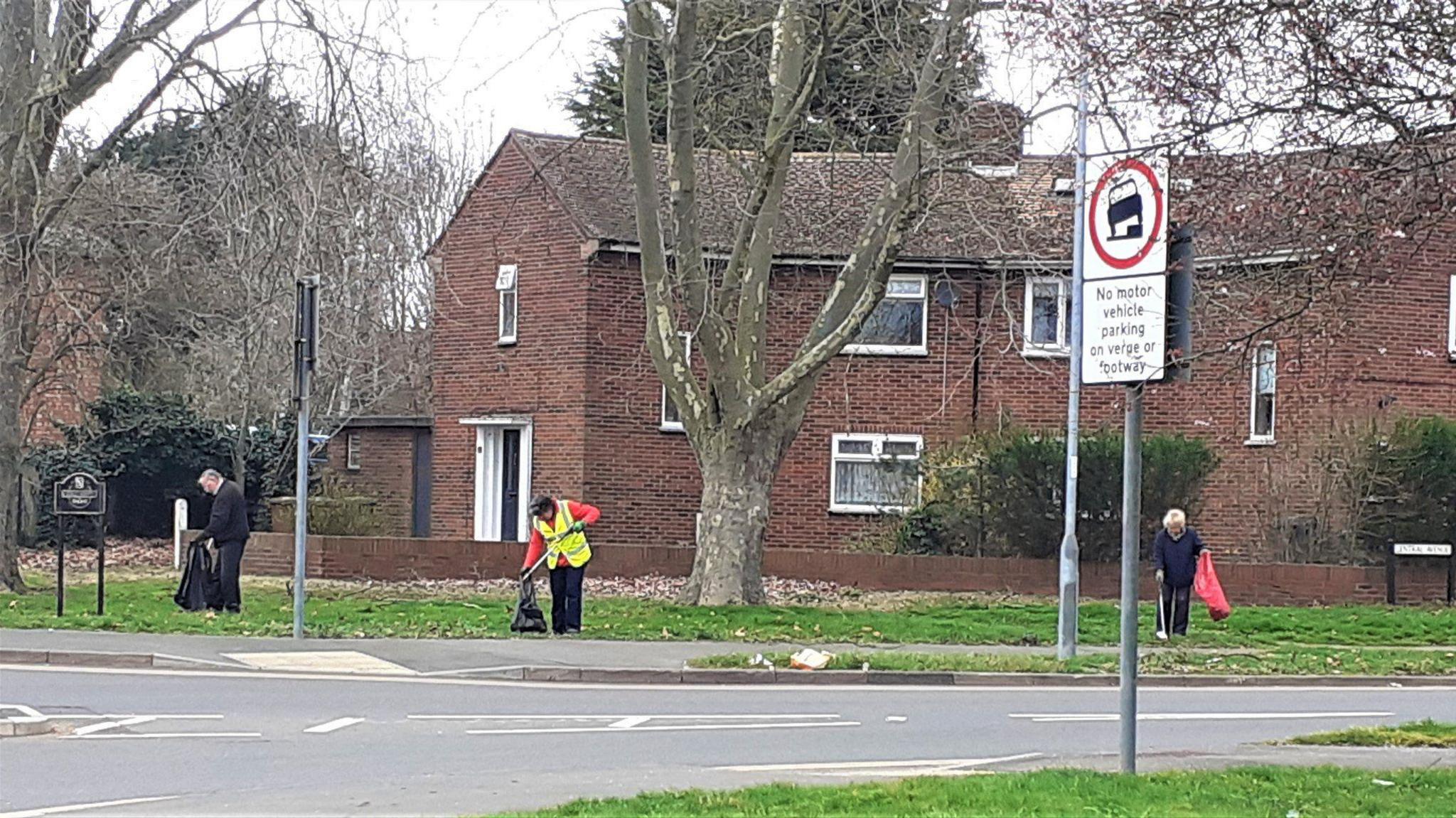 Three people collecting rubbish and litter in a residential street in Peterborough. There is housing behind them and grass verges and trees near the road junction. One person is wearing a high-visibility yellow tabard and the others are in dark clothing. 