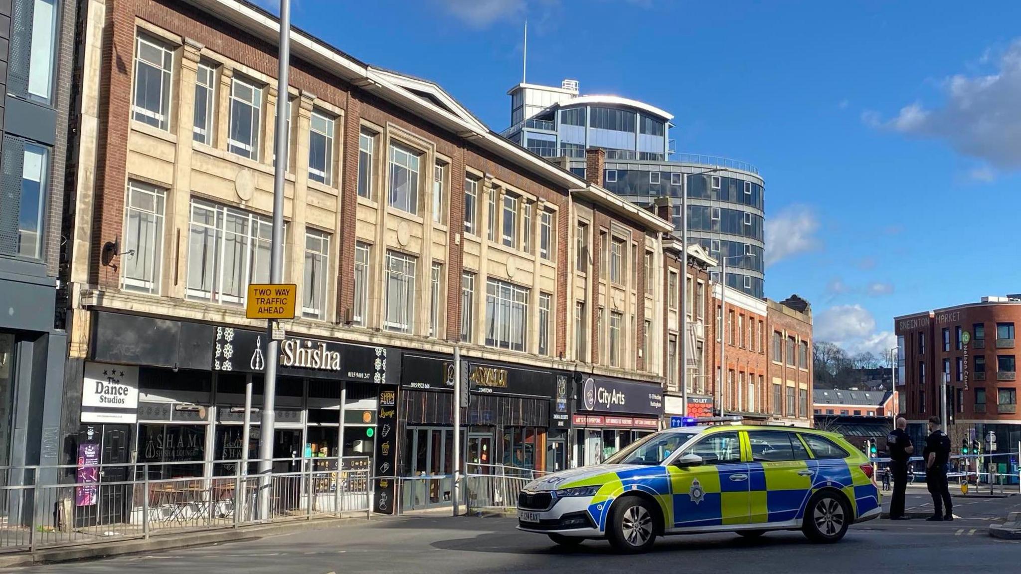 A police car is parked across a road. Two police officers in black clothes stand behind the vehicle looking away from the camera, police tape is behind them.