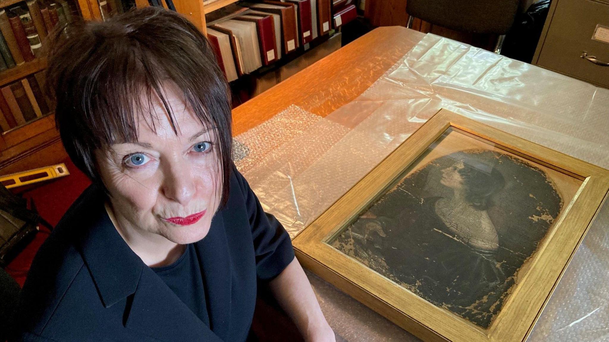 A woman dressed in black looking towards the camera with a painting in a gold frame on a wooden table behind her.