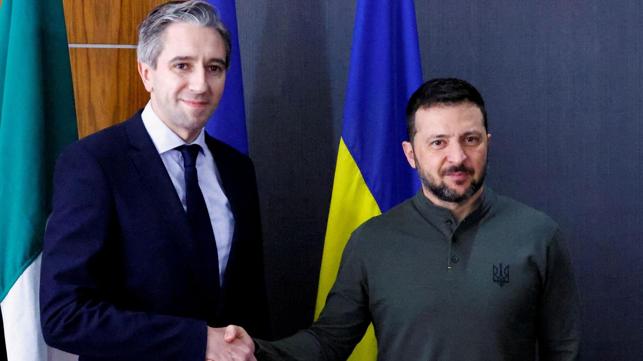 Simon Harris in a dark suit shakes hands with Volodymyr Zelensky who is wearing a green jumper. The Irish tricolour and Ukrainian flag are in the background.