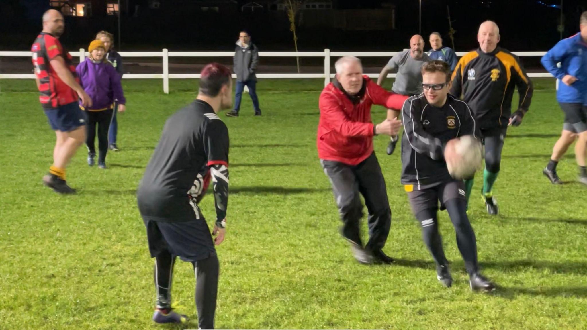 Ten rugby players can be seen on a floodlit pitch all wearing different strips and tracksuits. One man runs with the ball wearing a dark tracksuit top, shorts and athletic black leggings. A man in a red jacket and black tracksuit bottoms holds his hands out attempting to tackle the player.