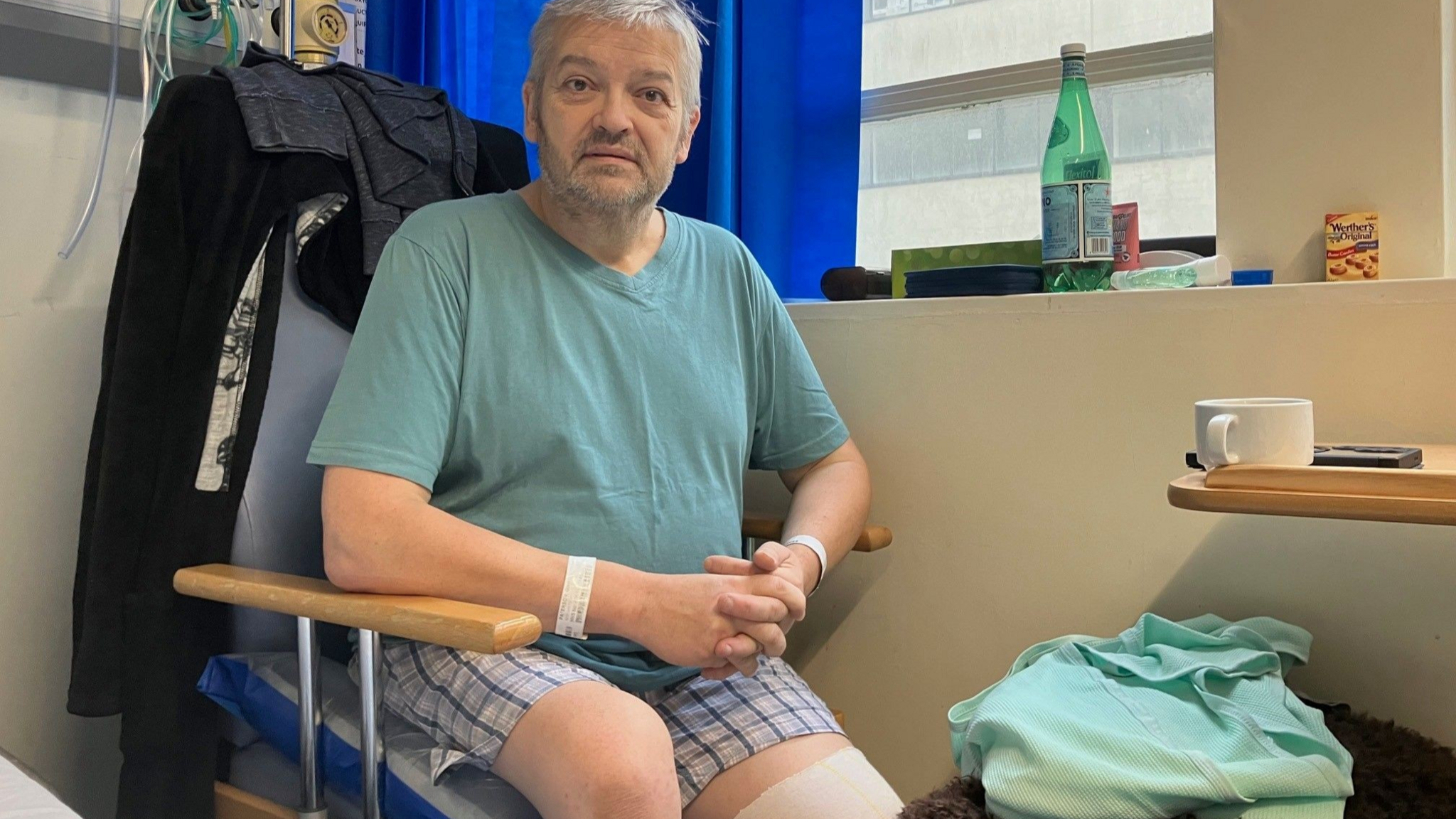 Glen Paterson sits in a hospital chair  wearing shorts, following an operation to remove his left leg below the knee. Hospital equipment is on the wall behind him and there are snacks and drinks on his windowsill