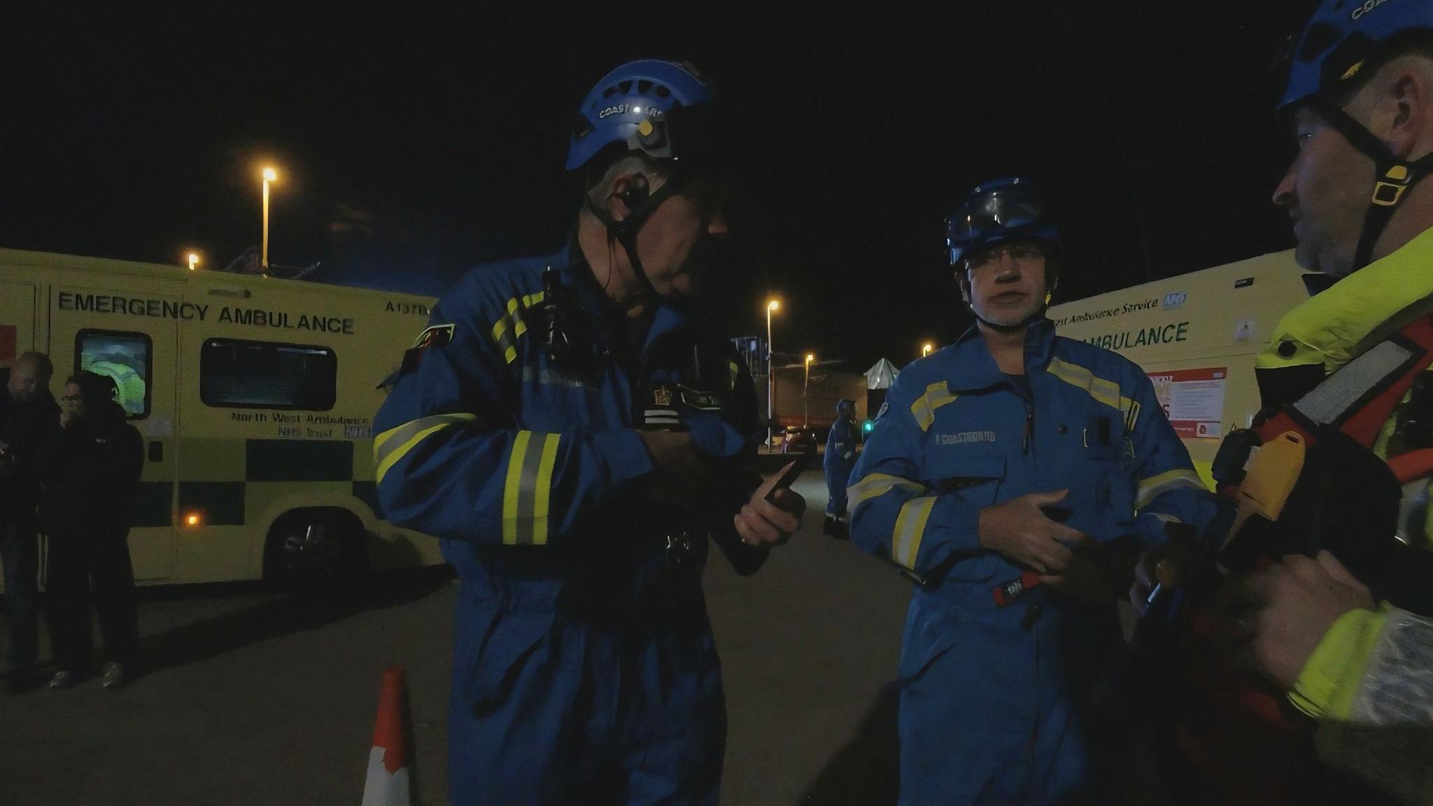 HM Coastguard rescuers discuss the plan during a major search. Two men in blue suits with yellow reflective patches stand in front of ambulances.