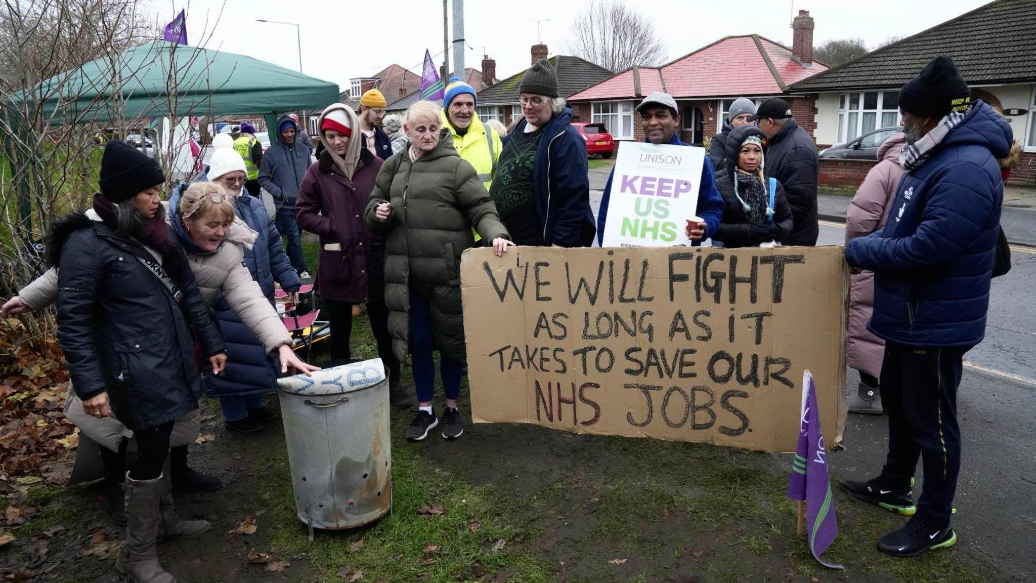 Men and women in warm coats outside on a pavement holding banners. One of the banners, written on cardboard, reads: "We will fight as long as it takes to save our NHS jobs."