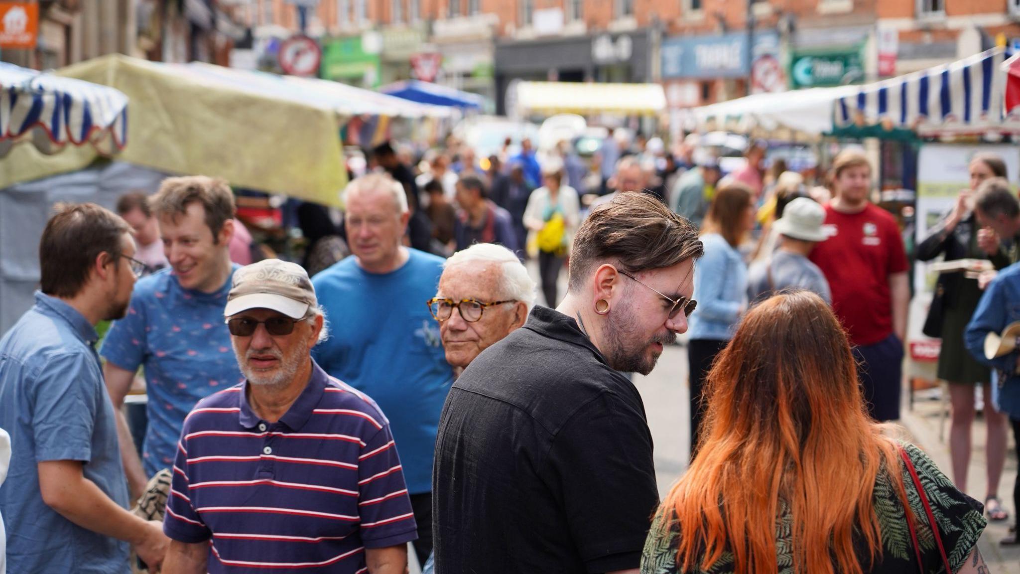 A general view image of the market with crowds of people walking around the market with stalls lining both sides of the street. 