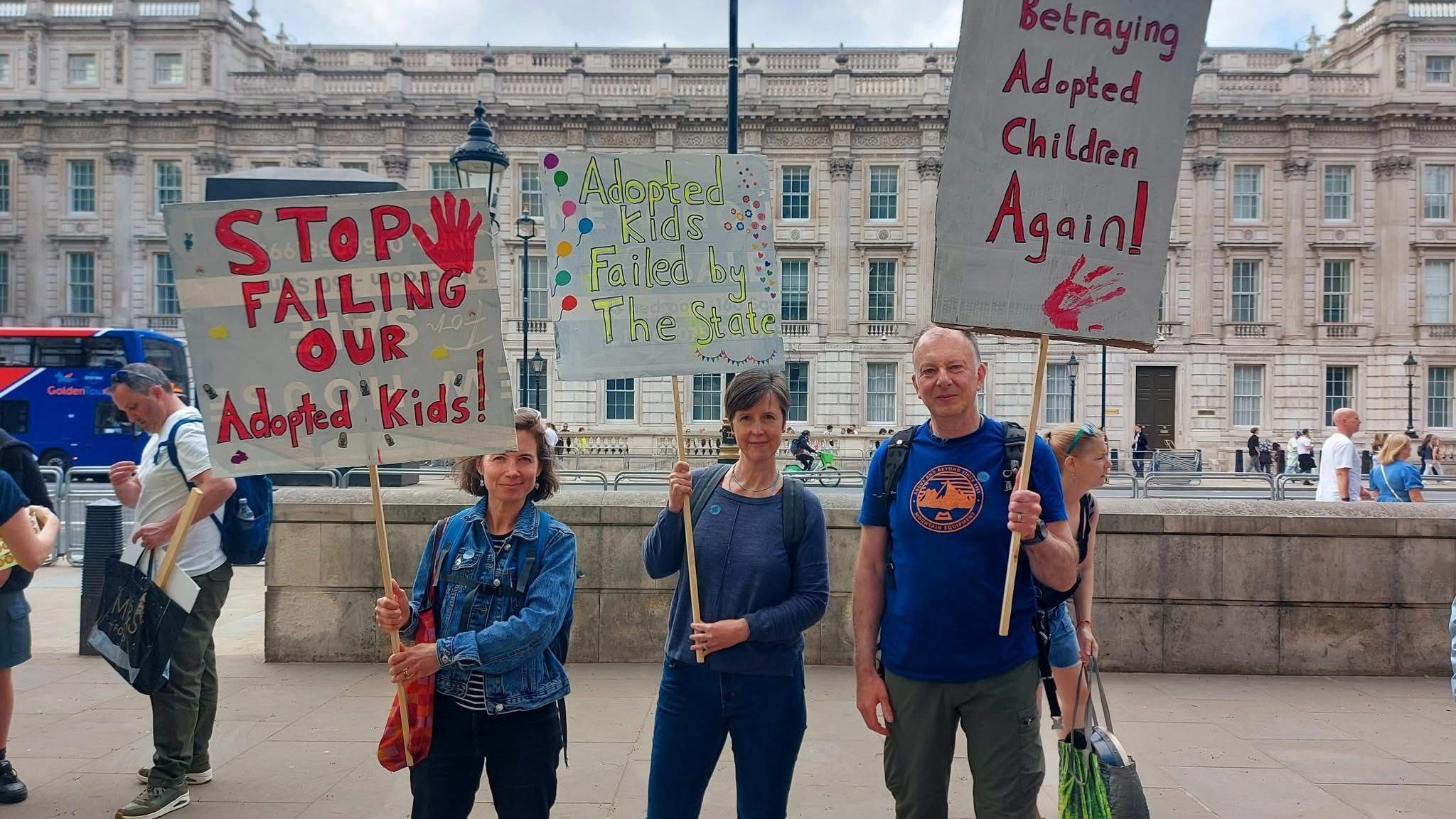 Three people all wearing blue hold placards above their heads. One reads 'stop failing our adopted kids' another reads 'betraying adopted children again'. In the background is a large stone building.