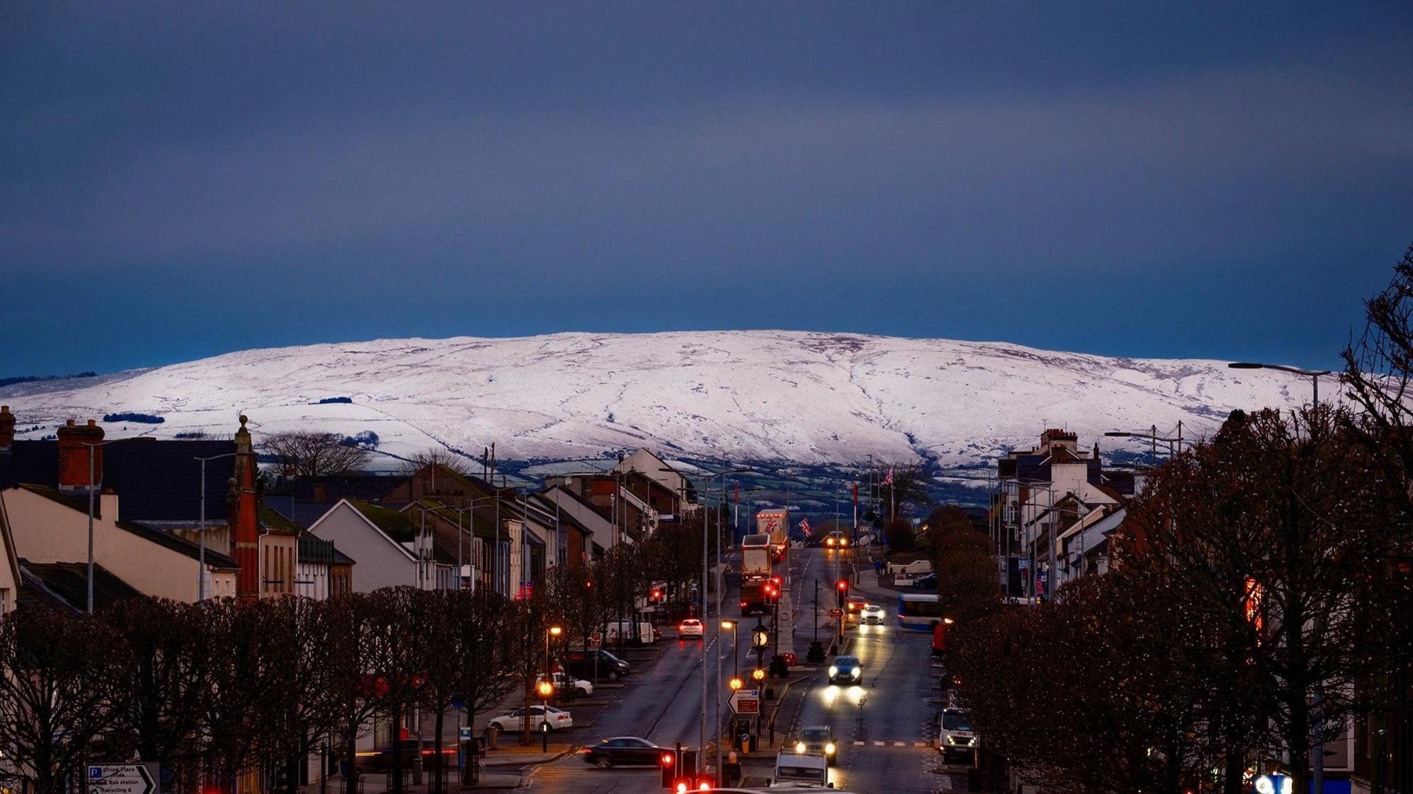 A white capped snowy hill sits above Cookstown main street, its evening and the road is busy with cars and buses