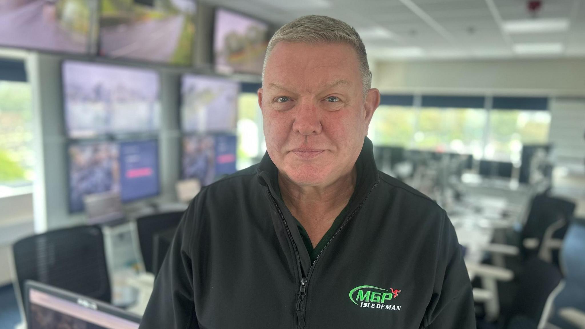 Clerk of the course Gary Thompson looking at the camera. He is wearing a black jacket with the MGP logo and standing in race control, which has large screens showing live videos of the TT course.