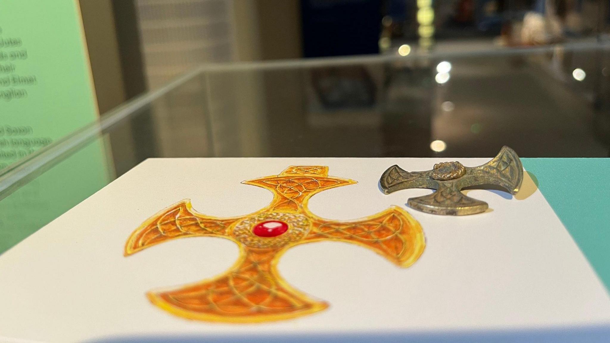 A small, ornate cross-shaped artefact with intricate patterns displayed on a white card. The card features a colourful illustration of the same cross design in gold with a red gemstone at the centre. The artefact is metallic and appears aged, while the illustration shows its original decorative appearance.