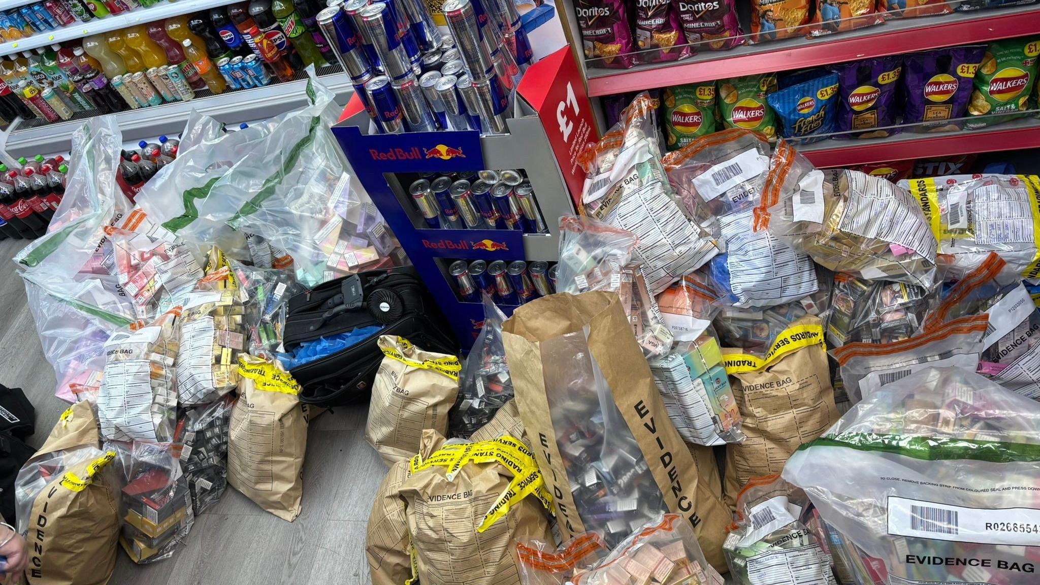 Clear bags of cigarette boxes and vapes can be seen on the floor in a newsagents. Behind the bags are shelves stocked with bottle fizzy drinks, bags of crips and a cardboard stand of Red Bull energy drinks. 