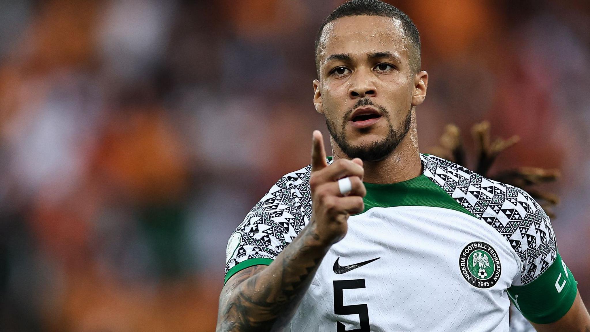 William Troost-Ekong, wearing a white Nigeria shirt with a tribal pattern on the shoulders, points a finger towards the camera as he celebrates a goal