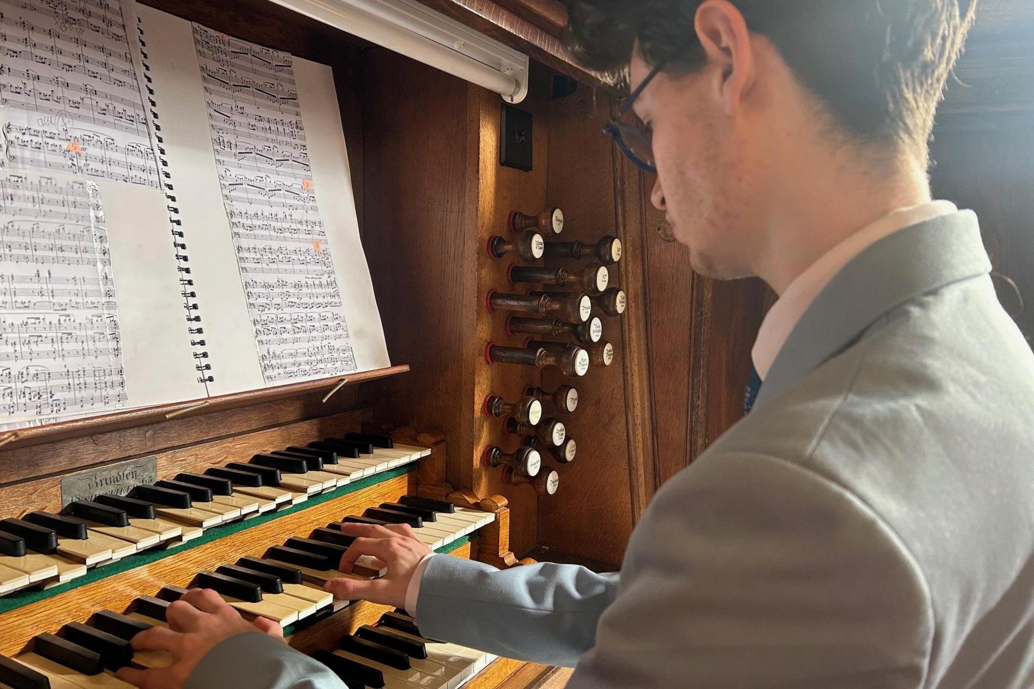 A close-up of Llewelyn Blezard-Greenwell playing the organ. He is playing the middle of three vertically stacked keyboards. Some of the keys on the top row are depressed and a bank of stops can be seen to his right and there is sheet music in front of him.