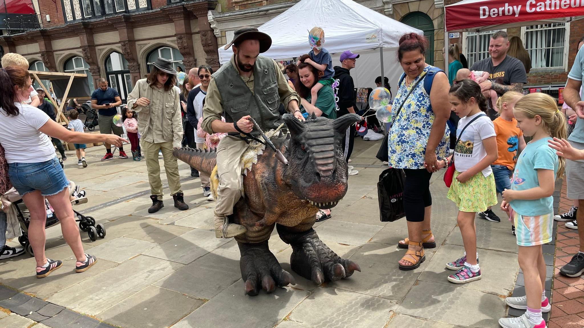 A man riding a plastic dinosaur while young children look on