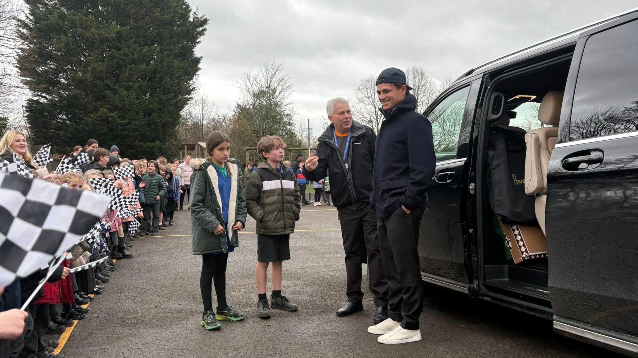 Photo of Lando Norris arriving at the school. He was great by all the children holding black and white checkered flags. He is standing next to a school teacher with his hands in his pocket. 