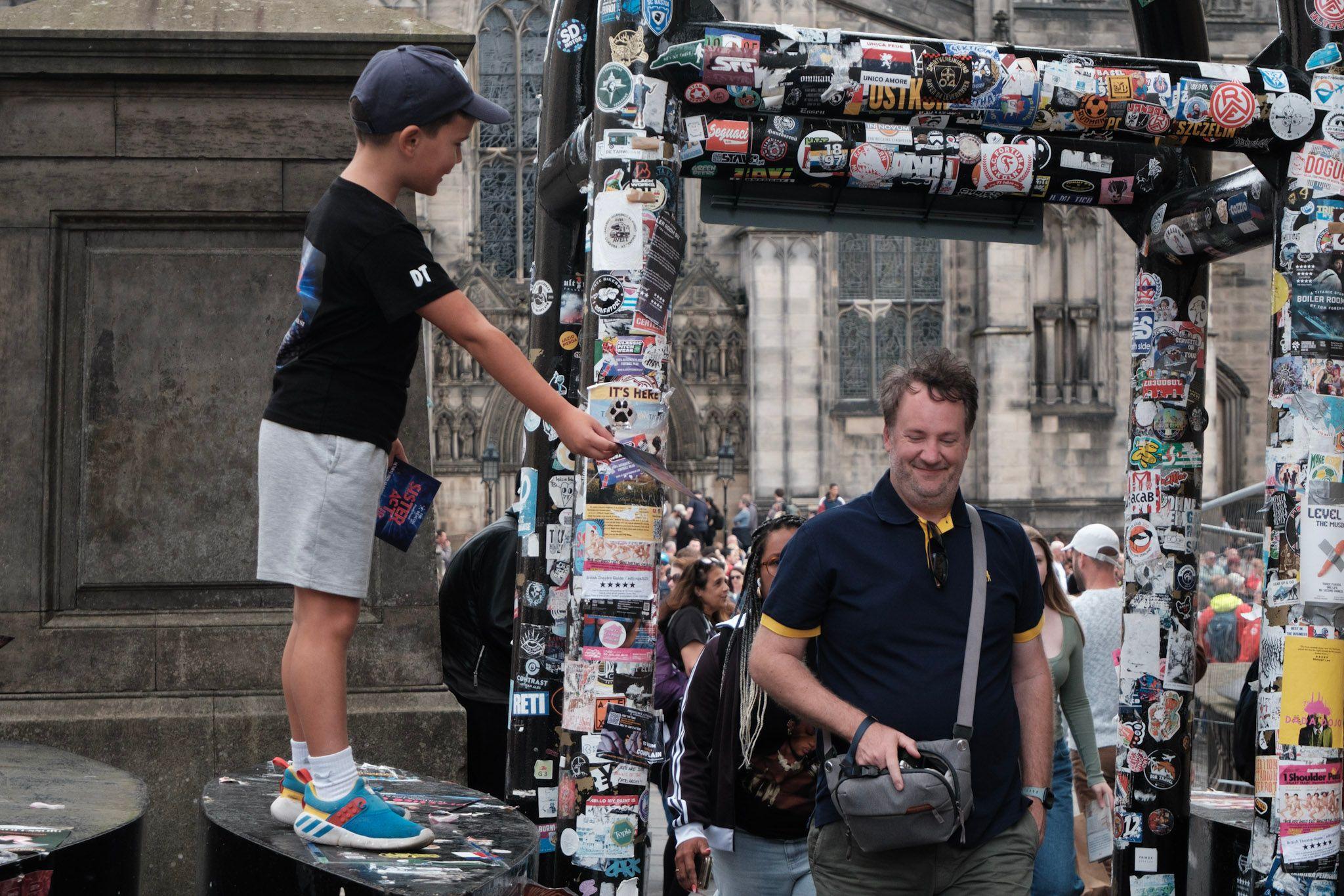 A young boy in shorts and a baseball cap gives out flyers standing on a street security bollard as people walk through the gate