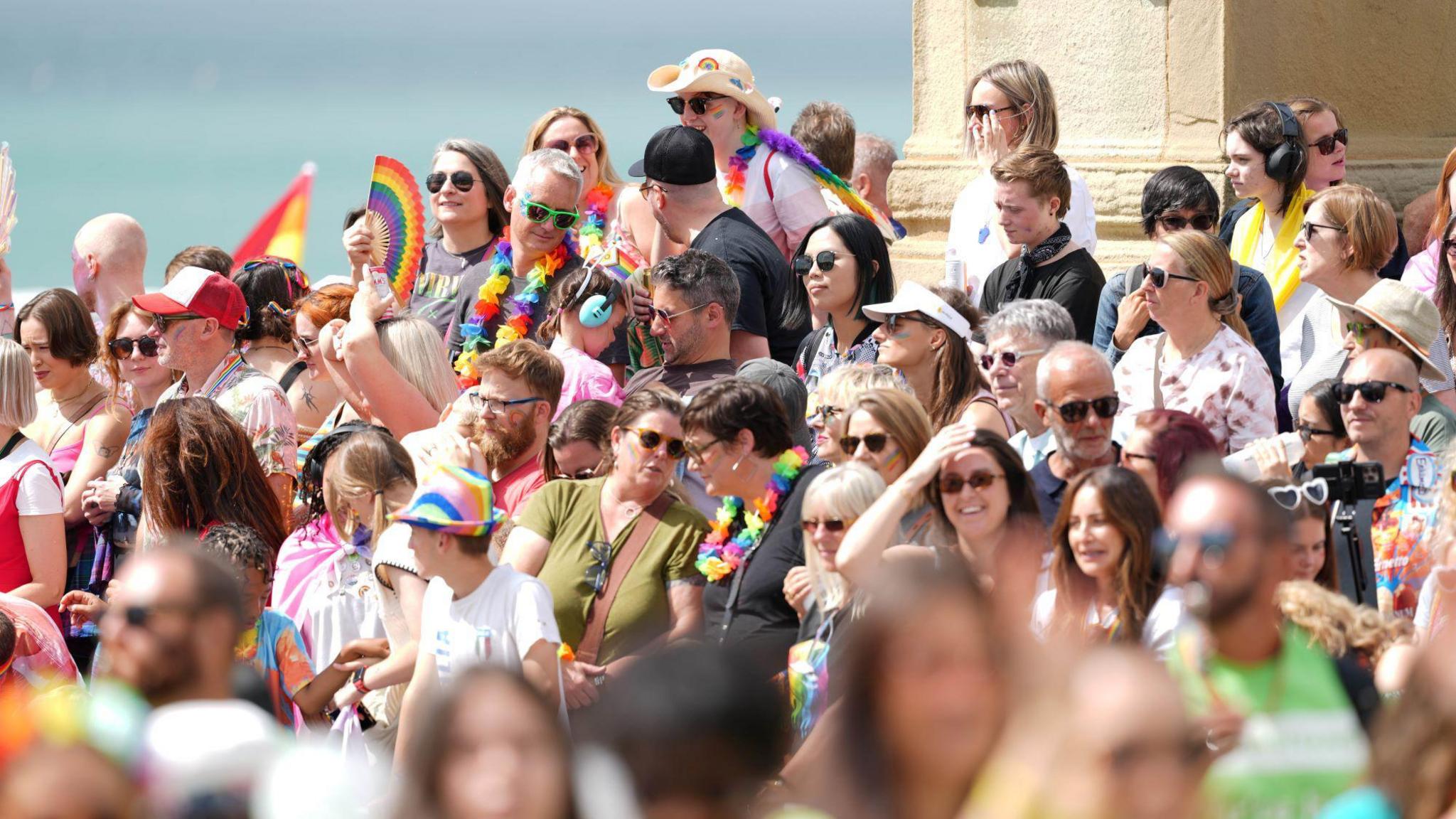 A crowd of people, some of whom are wearing rainbow hats and holding rainbow flags.