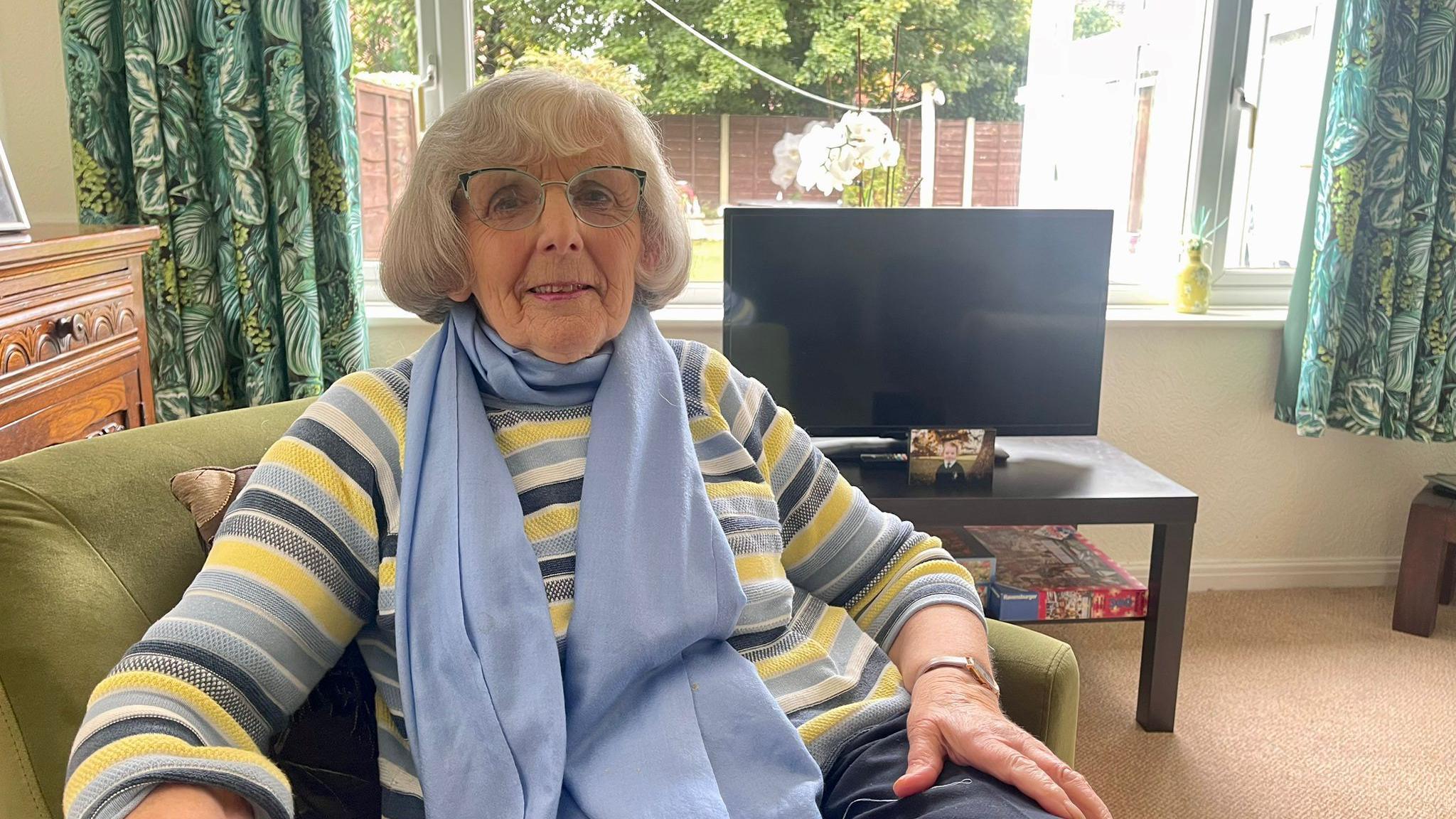 A woman with grey hair, glasses, a blue scarf and a striped jumper sits in a green armchair in her living room. Behind her is a flat screen television on a unit, a wooden chest and green floral patterned curtains that are open.