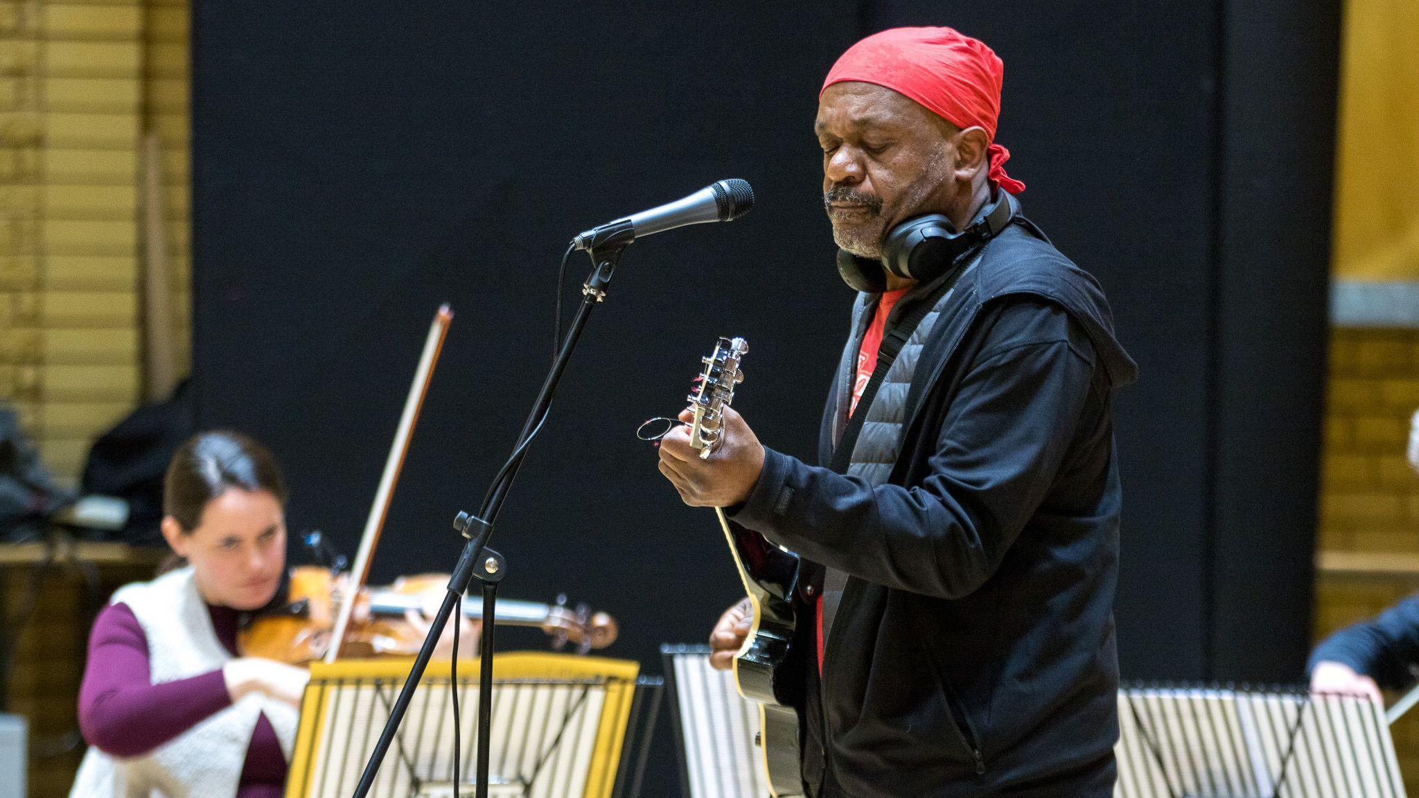 A man with a red head covering and headphones stands holding a guitar in front of a microphone. A woman sits in the back playing a violin.