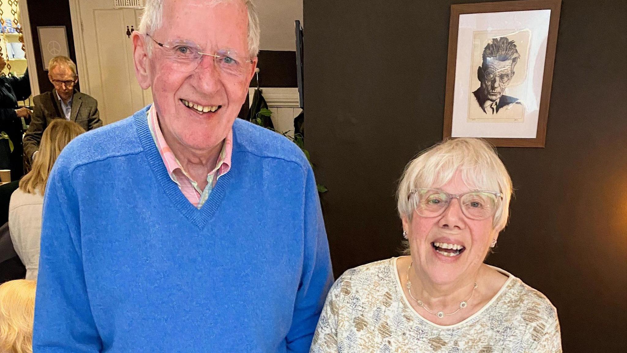 Two people standing indoors against a dark wall with a framed monochrome portrait hanging behind them. The person on the left is wearing a bright blue sweater over a collared shirt, while the person on the right is wearing a light patterned top.