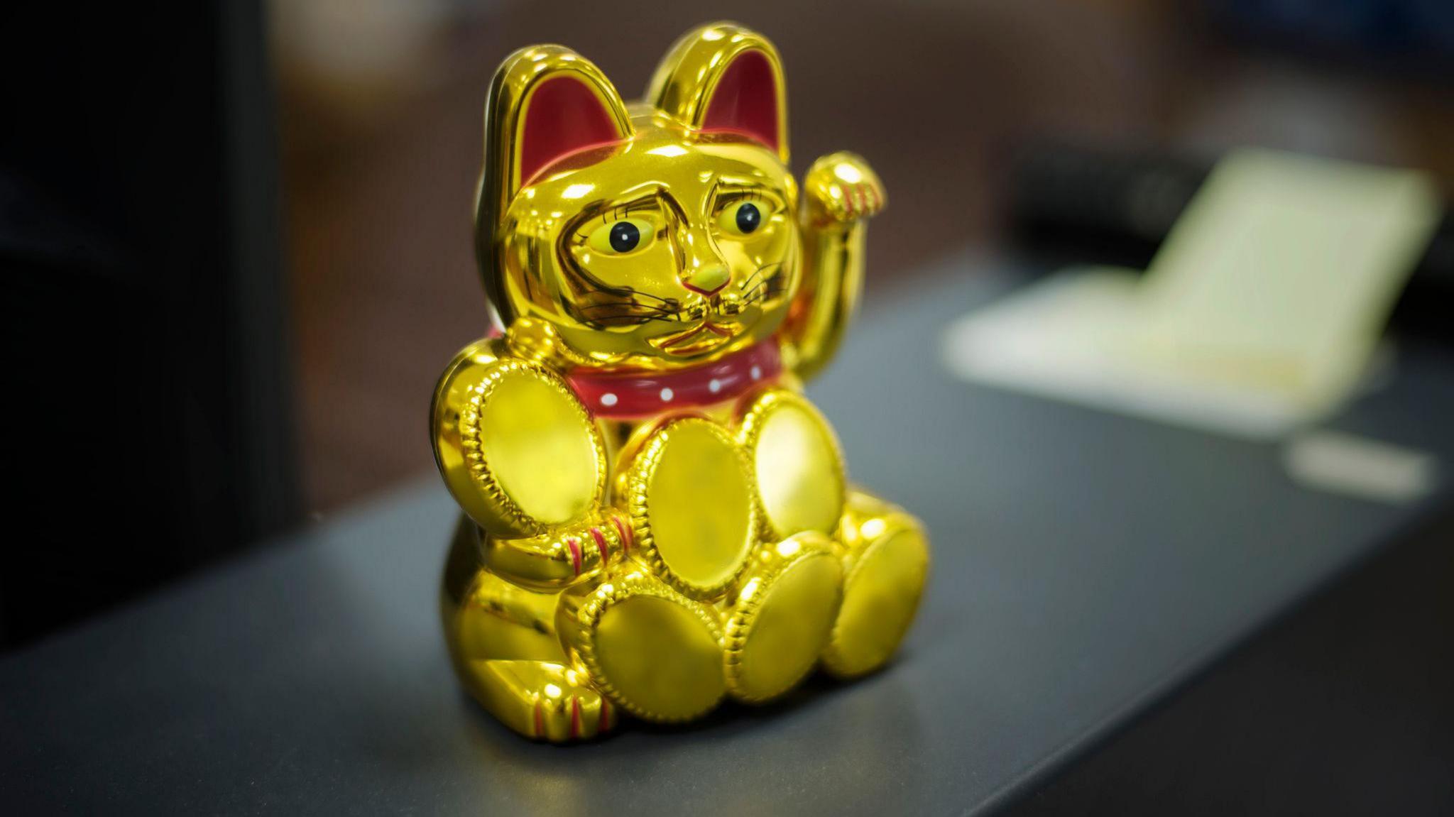 A shiny gold maneki-neko (beckoning cat) figurine with a red collar sits on a dark surface. The cat has a raised paw, large round eyes, and an exaggerated, slightly distorted face. The background is blurred, showing office-like elements.