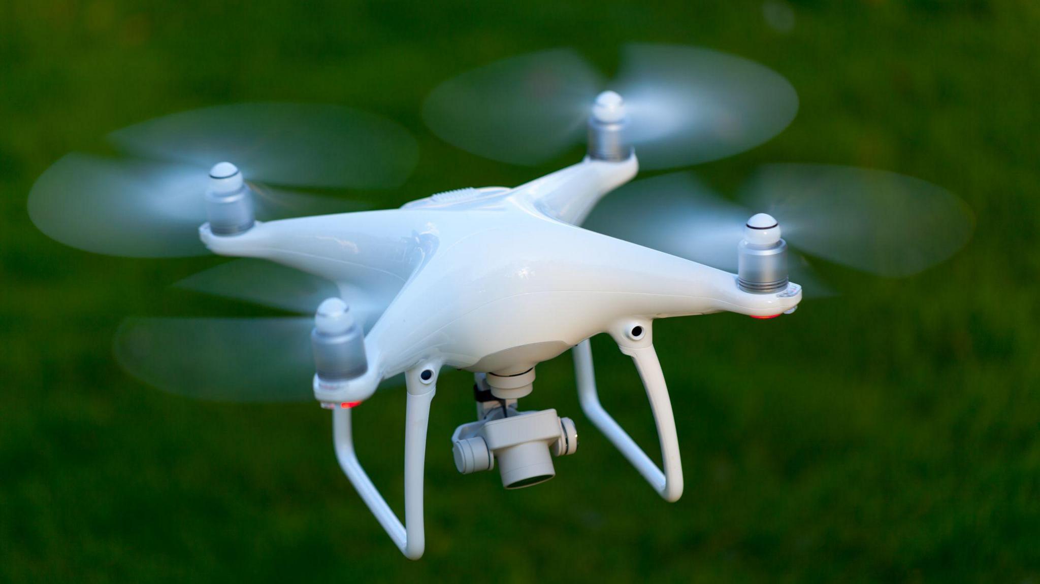 A white drone with four blades and a camera flies above the green countryside