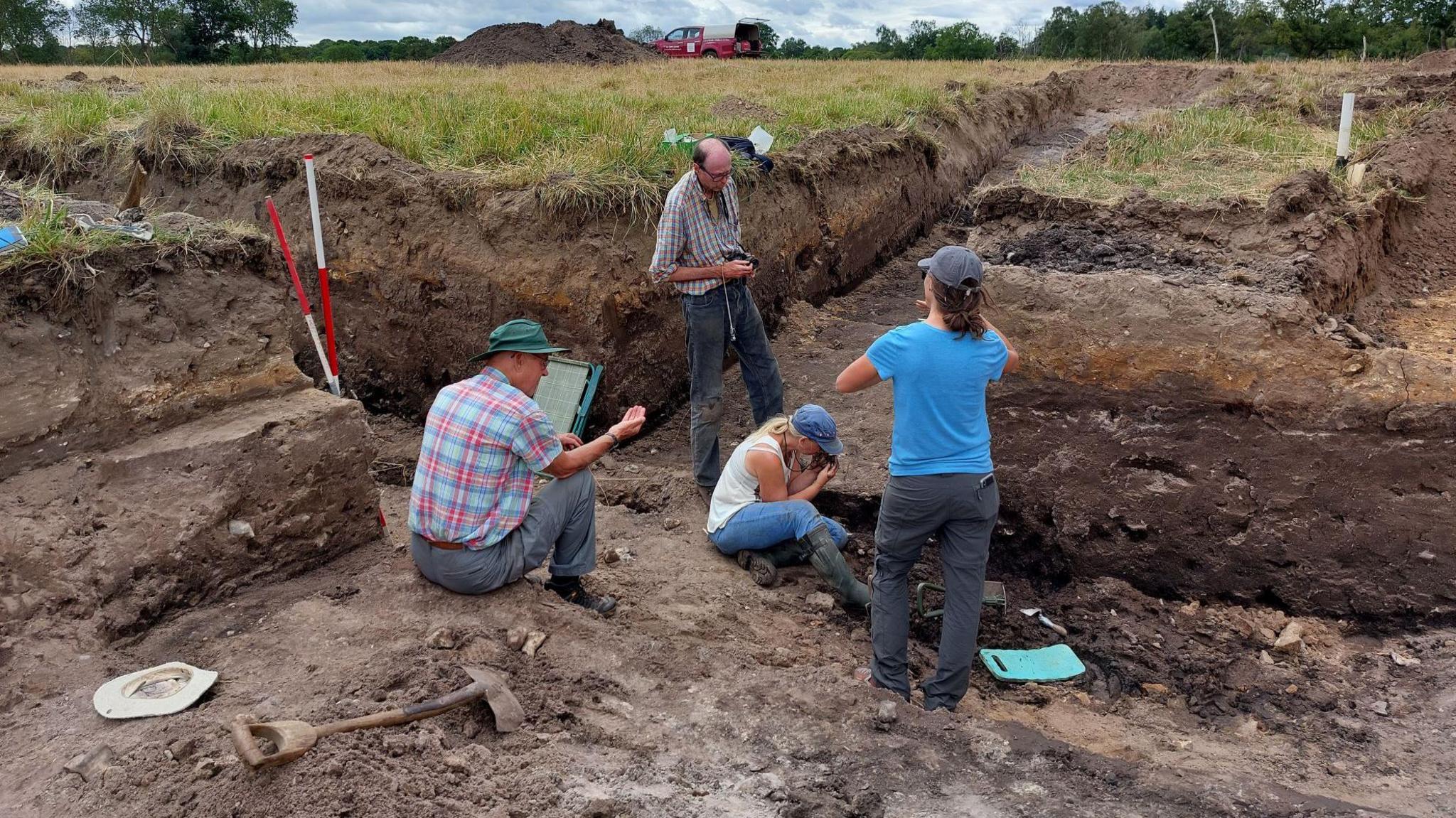 Men and women taking part in a dig in a field to excavate an ancient Ice Age pond. They stand in deep trenches at least five feet high (150cms). A truck and a mound of soil can be seen in the distance.