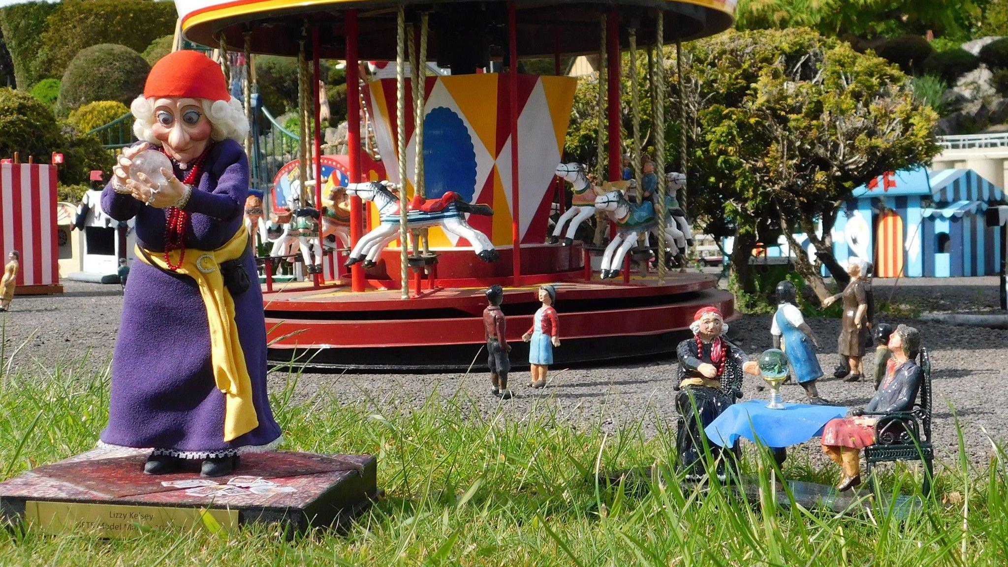A view of a fairground in a model village. A larger puppet of a fortune teller holding a crystal ball is stood close to where a miniature version of the same character is giving a reading as part of the model village.
